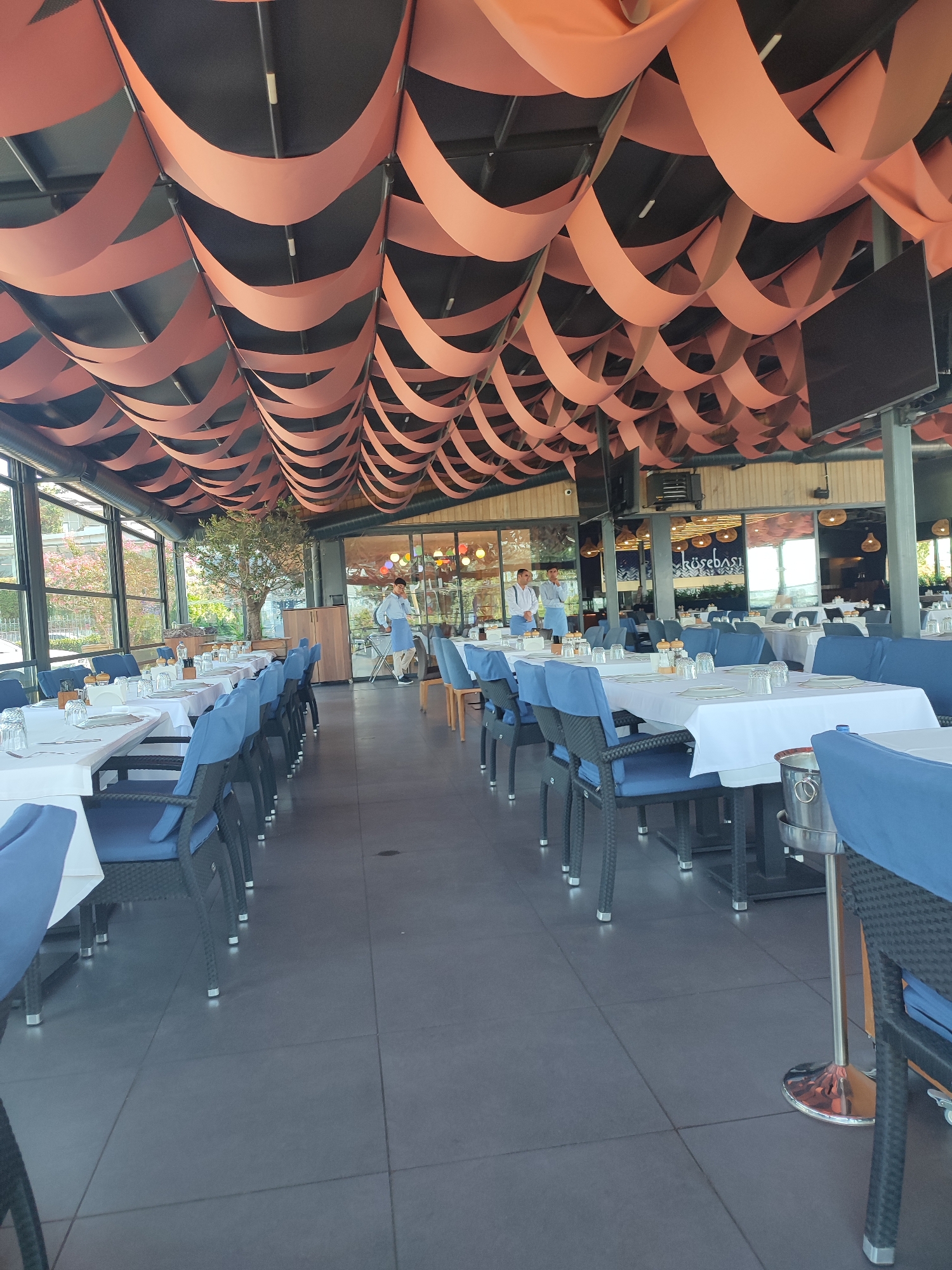 The image shows the interior of an outdoor seating area of a restaurant. The restaurant is located in the city of Üsküdar, Turkey. The ceiling is decorated with a series of orange fabric waves. The floor is tiled with dark grey square tiles. The tables are set with white tablecloths, white plates, and glasses. The chairs are black with blue cushions. There are two servers in the background, wearing blue aprons over white shirts and pants. One server is pushing a cart. The restaurant appears to be clean and well-maintained, with a modern design.