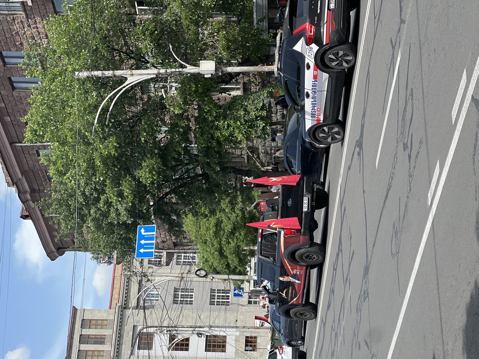 The image depicts a street scene in Yerevan, Armenia. A convoy of vehicles, including at least one Ford pickup truck, a black sedan, and a black Volvo SUV, is parked along the right side of a street marked with white lane dividers. The pickup truck is decorated with several small red flags, and it features a graphic of a person with a hand gesture, likely related to a political or social movement. The Volvo SUV is marked with the word "POLICE" in Armenian script (ՈՍՏԻԿԱՆՈՒԹՅՈՒՆ), along with the numbers 112 and 1256, indicating it is a police vehicle.
The street is lined with buildings, including older stone structures and a multi-story grey building with numerous windows. A blue traffic sign with white arrows indicates the permitted direction of travel for different lanes. Overhead power lines and a vintage-style lamppost with curved arms are visible. The presence of a crowd of people further down the street suggests a gathering or procession.