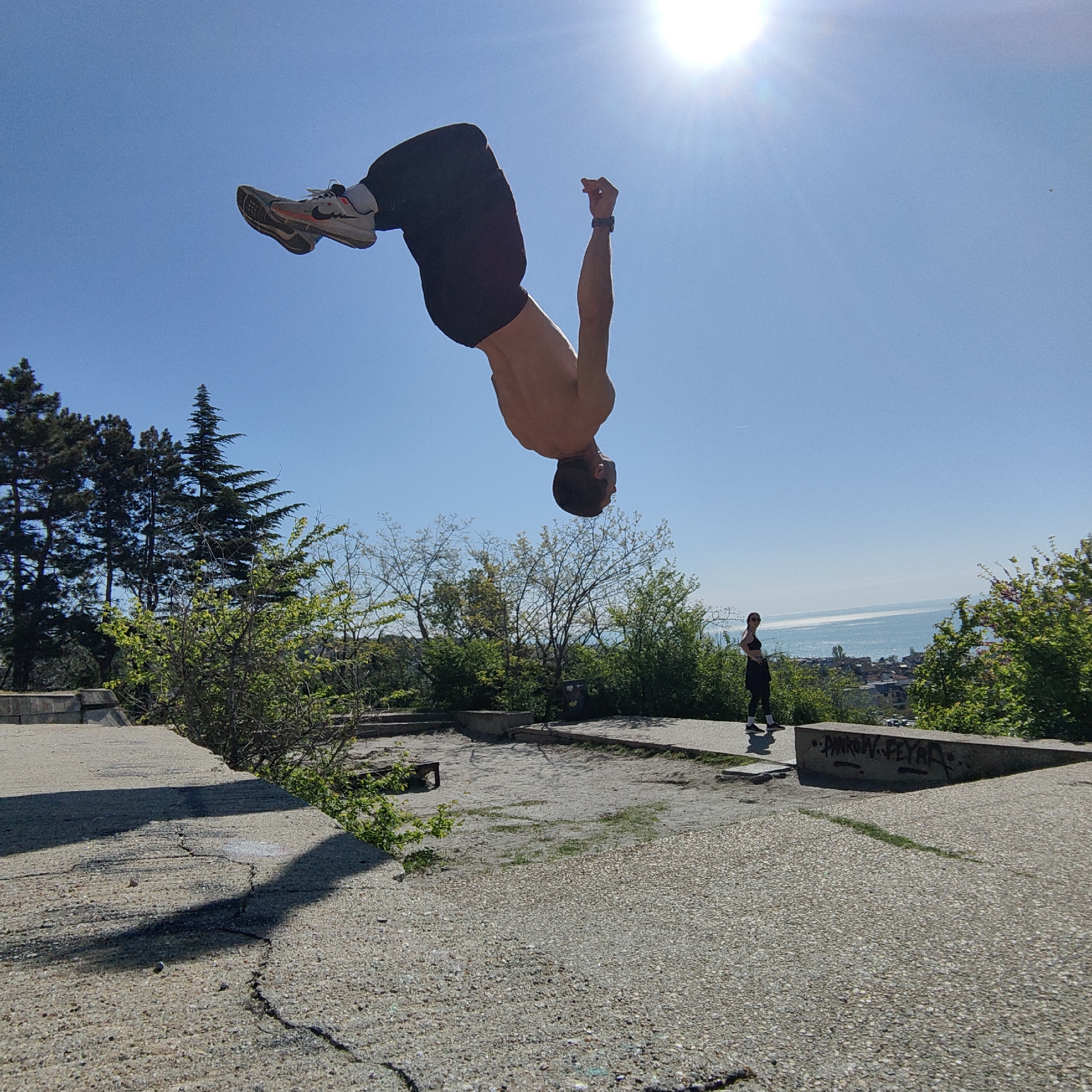 The image shows a shirtless man performing a backflip in Varna, Bulgaria. He is wearing black pants and Nike shoes. The background includes trees, a sea view, and another person standing on a concrete platform. The concrete surface is cracked. The sky is bright blue with the sun visible.