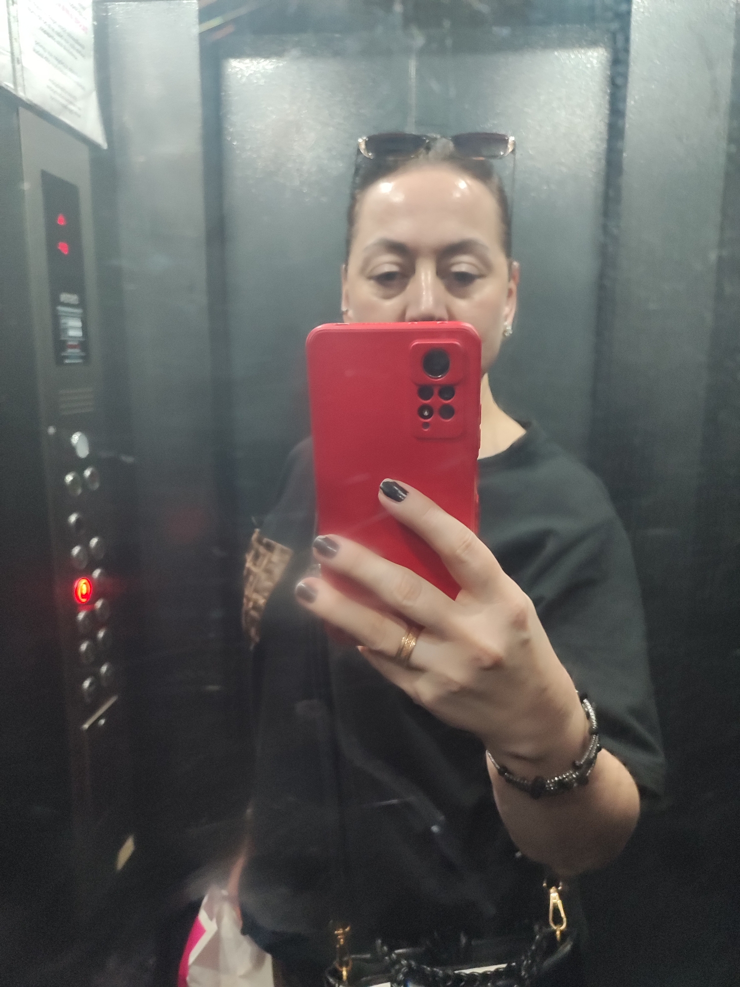 A woman is taking a selfie in an elevator. She is holding a red smartphone in her right hand, with her left hand resting on her hip. She is wearing a black t-shirt, sunglasses perched on her head, and a gold ring on her left ring finger. She also has on a bracelet with black beads. The elevator's control panel, with illuminated floor numbers, is visible to her left. The elevator walls appear to be made of reflective metal. The overall scene is dimly lit.