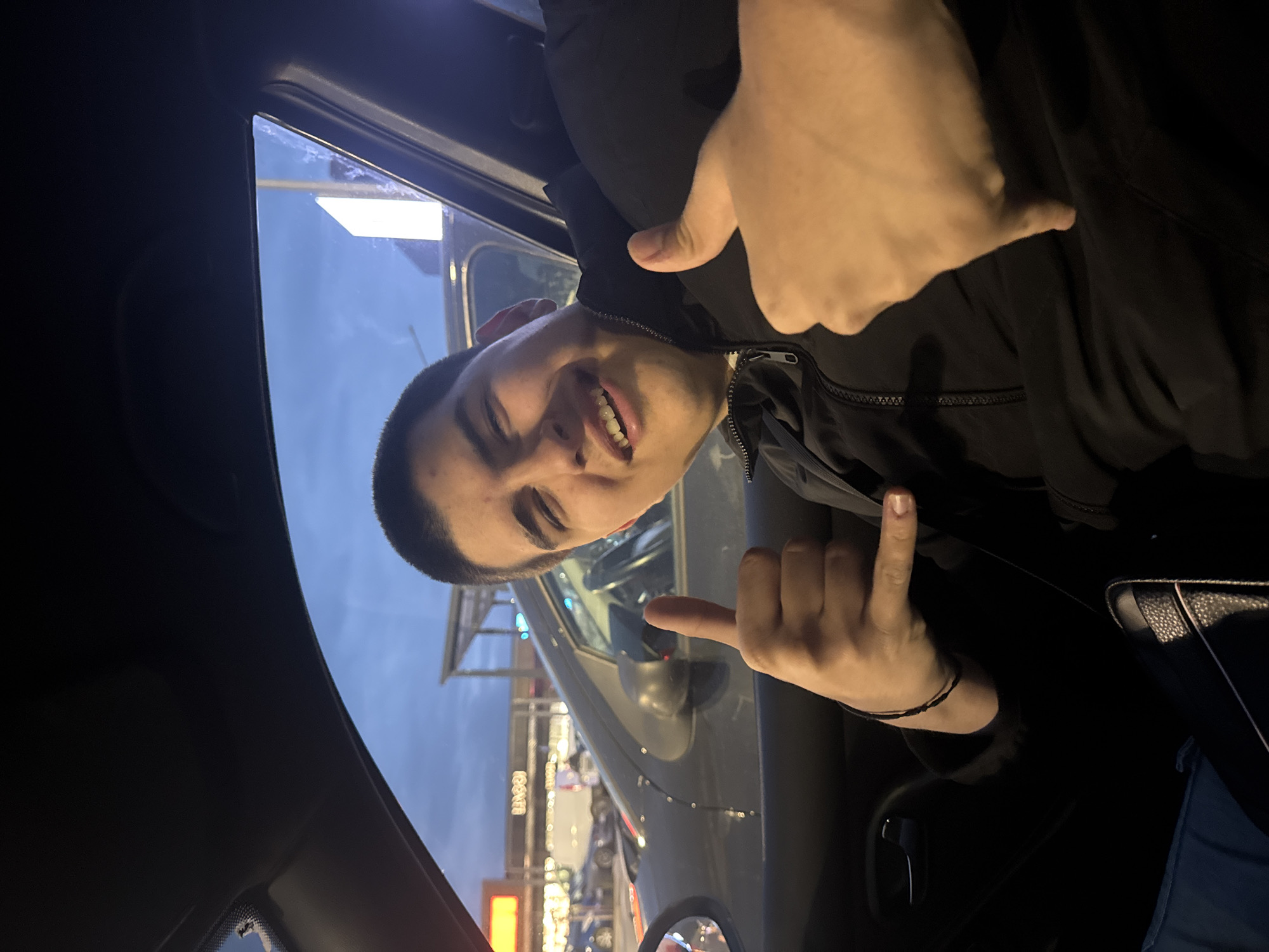 A young man is in the driver's seat of a car, smiling and gesturing with his hands. He is wearing a black jacket. Through the car window, storefronts and signs are visible, including a sign that reads "ТЕХНОПОЛИС" (TECHNOPOLIS), which is a major electronics retailer in Bulgaria. The location is likely in Sofia, Bulgaria. It is dusk or evening, judging by the sky and the artificial lighting.