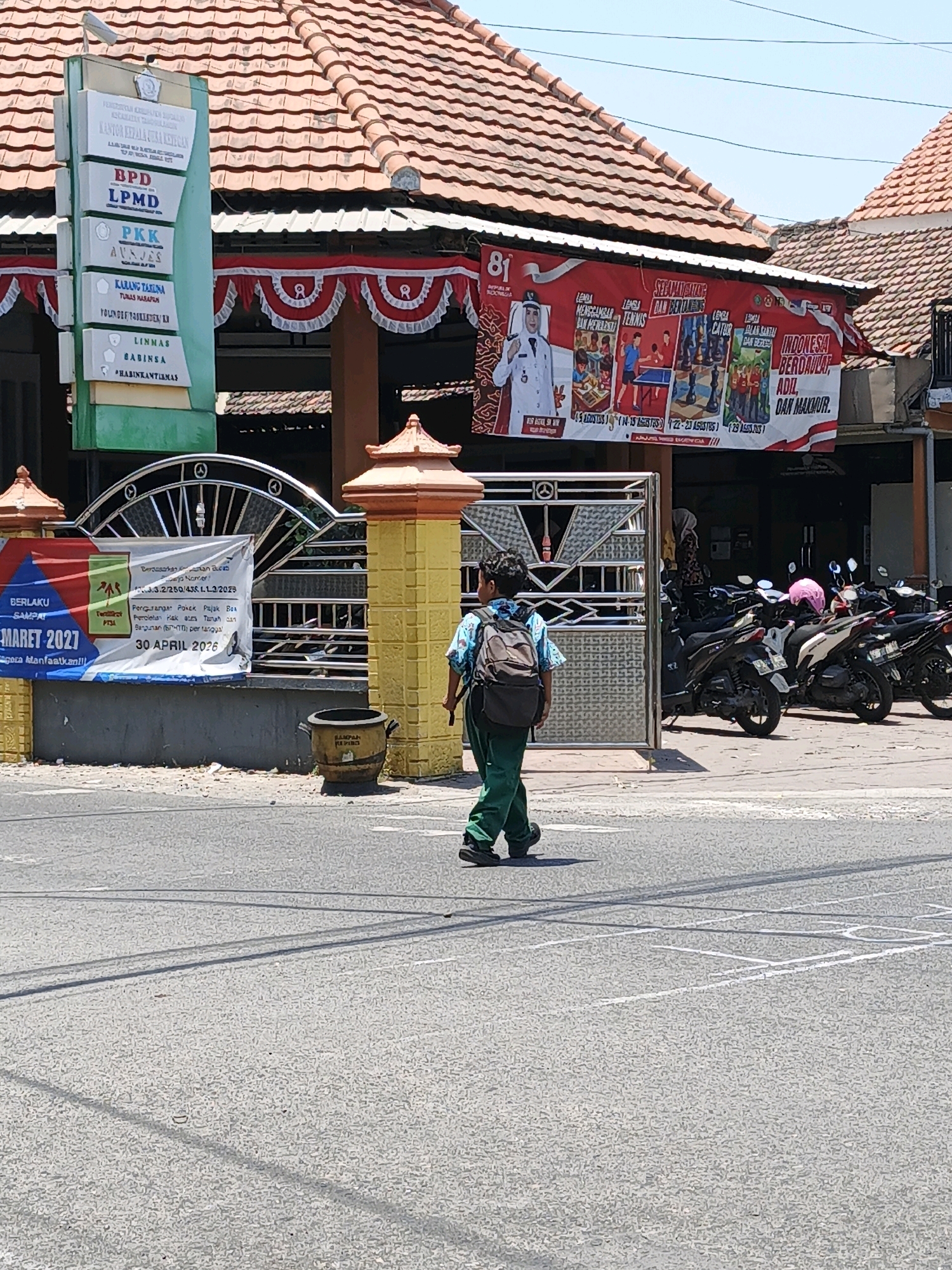A boy wearing a green uniform and a backpack walks on a street in front of a building with a red tiled roof. The building has a sign with the text "Kantor Kepala Desa Ketegan" (Ketegan Village Head Office). A banner with Indonesian text and images is hung above the entrance. Several motorcycles are parked in front of the building. The street is paved and has white markings. The sky is clear and blue.