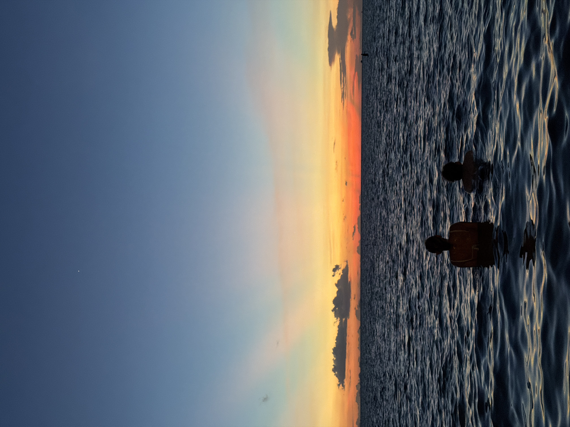 The image depicts two individuals swimming in the ocean at dusk, with the horizon dominated by a vibrant sunset. The sky transitions from a clear blue at the top to shades of orange and yellow near the horizon, punctuated by scattered clouds. The ocean's surface is characterized by dark, rippling waves. In the foreground, two silhouettes are visible in the water, with their heads and upper bodies exposed. The one on the left appears to be wearing a bathing suit, while the other on the right is also partially submerged. The scene suggests a serene and tranquil atmosphere. There is a single, small, bright point in the upper part of the sky, possibly a planet or star. No specific landmarks in Baan Plai Laem, Thailand are identifiable.