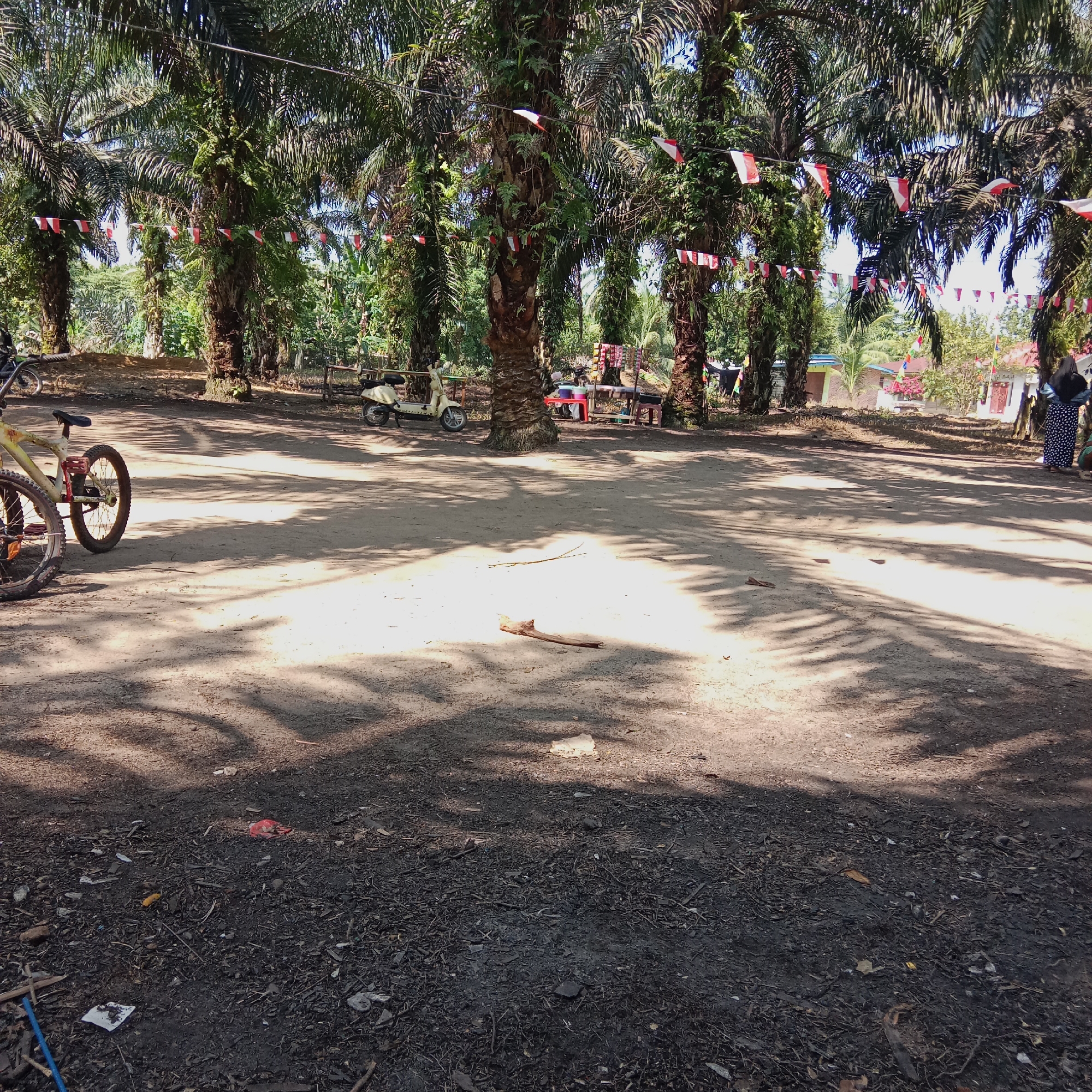 The image depicts an outdoor scene, likely in Dumai City, Indonesia, characterized by a dirt ground area with scattered debris. In the foreground, two bicycles are partially visible, suggesting they are parked or abandoned. The background features a row of palm trees, many adorned with small red and white flags strung between them, indicating a possible celebration or national event. Beyond the palm trees, there are buildings and more vegetation. A light-colored scooter is parked to the left of the scene, and in the mid-ground, a small market stall or setup is visible with a red table and some items on display. Several individuals are present in the background, with one person on the far right wearing a dark headscarf and patterned skirt, and another person partially visible in the lower right corner. The lighting suggests it is daytime, with dappled sunlight casting shadows from the palm fronds onto the ground.