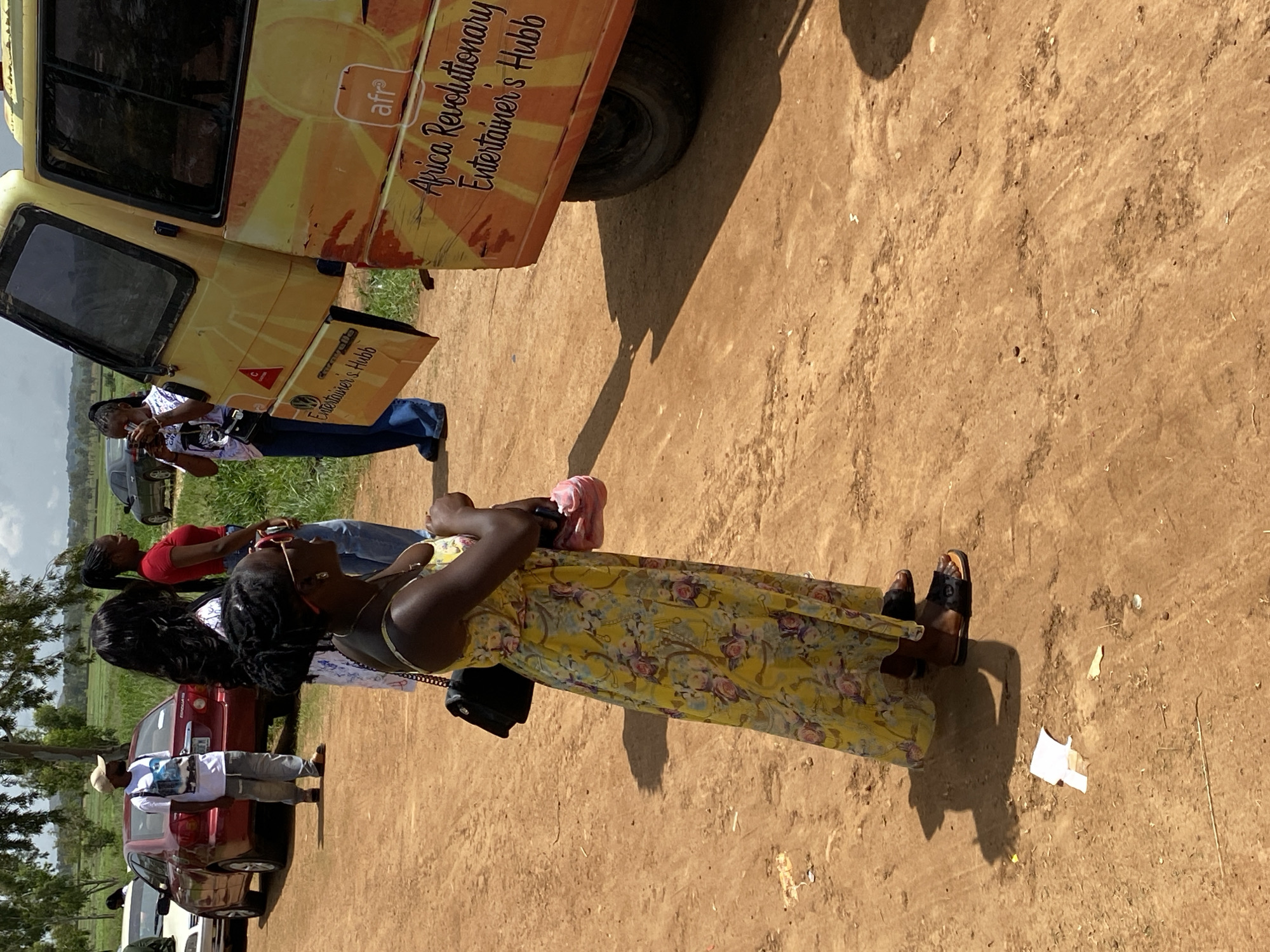 In Durumi, Nigeria, a group of individuals is gathered outdoors on a dirt road. In the foreground, a woman wearing a yellow floral print dress and sunglasses stands facing a yellow van with the words "Africa Revolutionary Entertainer's Hubb" written on its side. She holds a pink object in her right hand and has a black purse slung over her left shoulder. Behind her, two other women are visible, one in a red shirt and the other with long dark hair. To the left, a man in a light-colored cap and white t-shirt stands near a red car. The environment is dusty with scattered vegetation in the background.