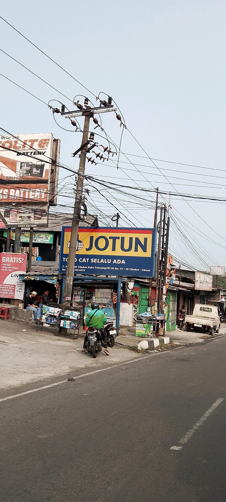 This image captures a street scene in Gunung Putri, Indonesia, featuring a prominent "JOTUN" sign and various roadside businesses. A utility pole with numerous wires dominates the center of the frame. On the left, a banner with "GRATIS!" and "CEK AKI" is visible, suggesting promotional activity. Several individuals are present: two men are seated on a ledge near a stall displaying what appear to be car batteries and other products, while another person in a green shirt is on a motorcycle. A white pickup truck is parked on the right side of the road. The environment is characterized by a paved road, a sidewalk or paved area in front of the shops, and overhead electrical lines. The sky is overcast and pale.