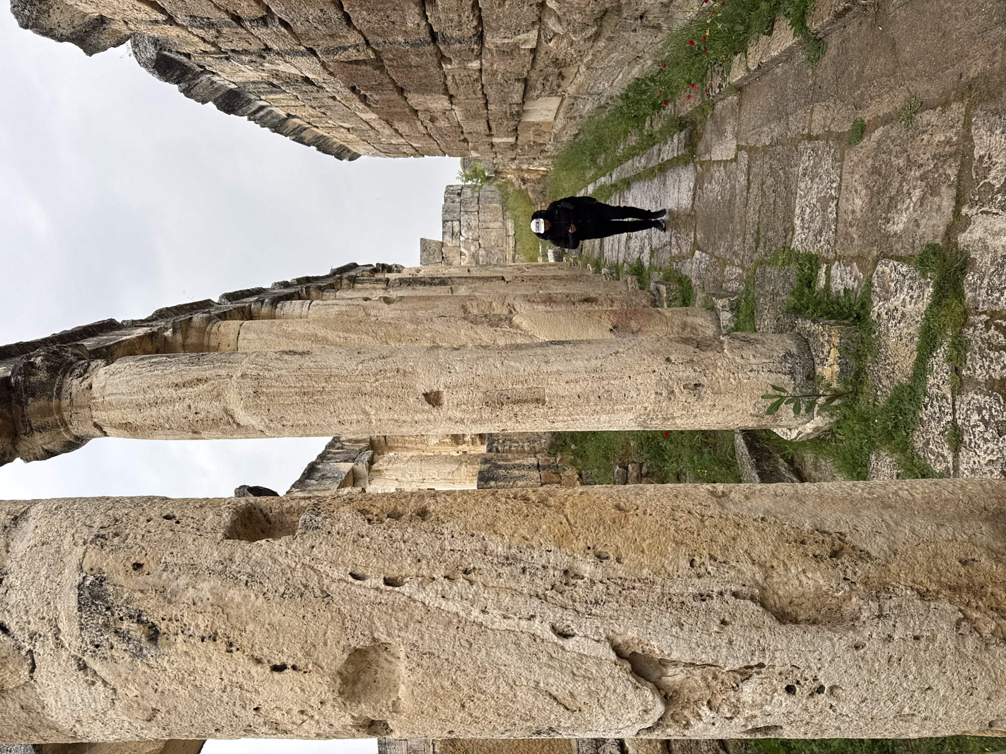 The image shows the ruins of Hierapolis, an ancient city located near Pamukkale, Turkey. A person dressed in black with a white face covering is walking along a stone pathway between tall stone walls and rows of columns. The structures are weathered and appear to be made of limestone. Vegetation, including green grass and some red flowers, grows along the edges of the pathway. The sky is overcast.