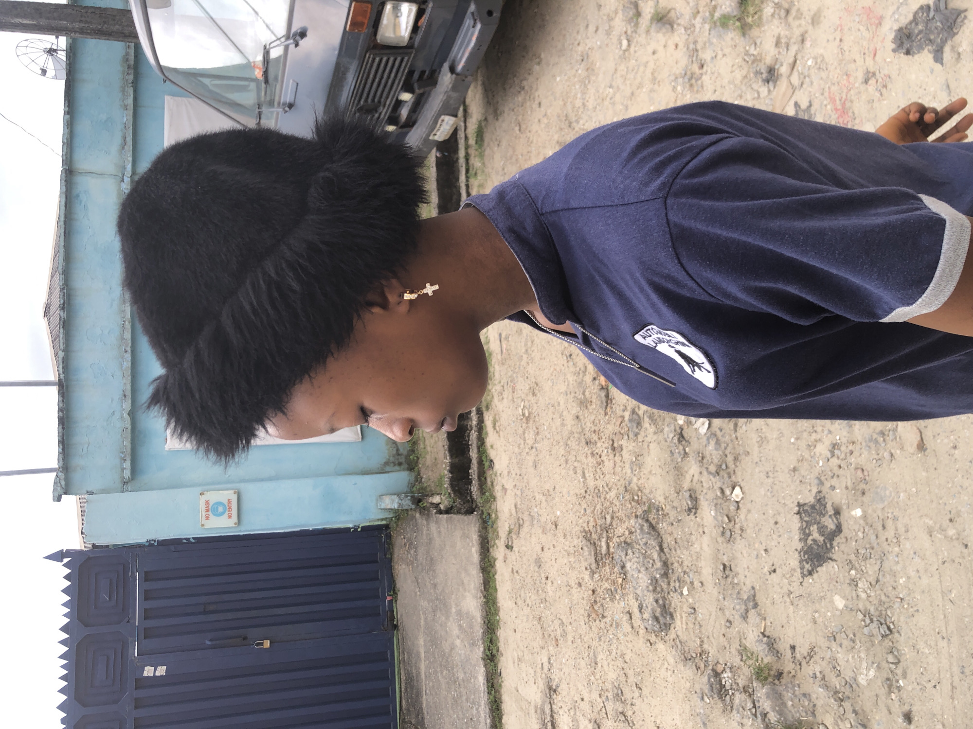 A young person wearing a black fuzzy hat, a navy blue polo shirt with a patch on the left breast, and a cross earring stands in an outdoor urban environment. The person is facing to the left with their eyes closed. Behind them is a light blue building, a dark blue metal gate with a padlock, and a dark-colored van. The ground is a mixture of dirt and gravel with some visible debris. A sign on the blue building reads "NO MASK" and "NO ENTRY". This scene appears to be in Ado, Nigeria.