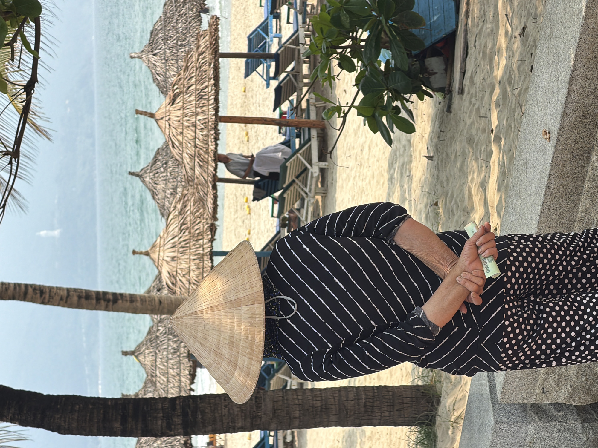 A daytime image captures a person on a beach in Đà Nẵng, Vietnam. The individual wears a traditional conical hat and a black and white patterned outfit. In their hands, they hold rolled Vietnamese currency. In the background, straw umbrellas line the beach, and beachgoers are visible on lounge chairs near the shoreline. A tall palm tree frames the left side of the frame, while a staircase and foliage are seen to the right. The sky is visible in the upper portion of the frame.