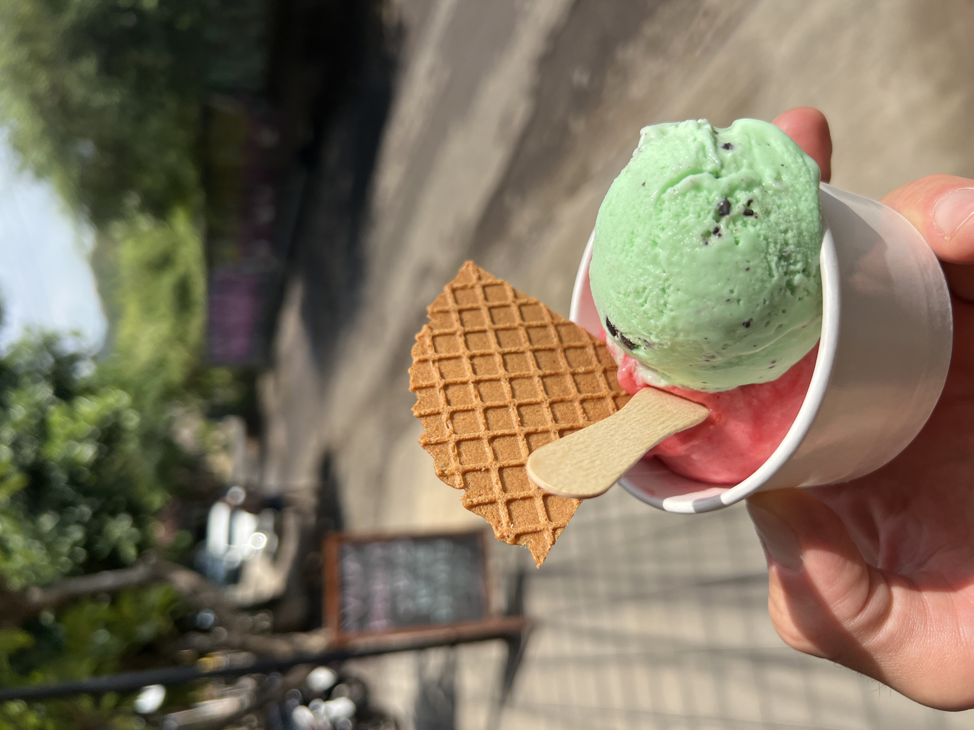 A person's hand holds a white paper cup filled with two scoops of gelato, one pink and one mint green with dark chocolate chips. A waffle cookie shard and a wooden spoon are inserted into the gelato. The background is blurred, showing a street scene with trees, a parked motorcycle, and a blackboard sign. The location appears to be a sidewalk in the city of Abang, Indonesia, based on the architectural and vegetative context suggesting a tropical environment with a relaxed, possibly tourist-oriented atmosphere. The lighting indicates it is daytime, with bright sunlight casting shadows.