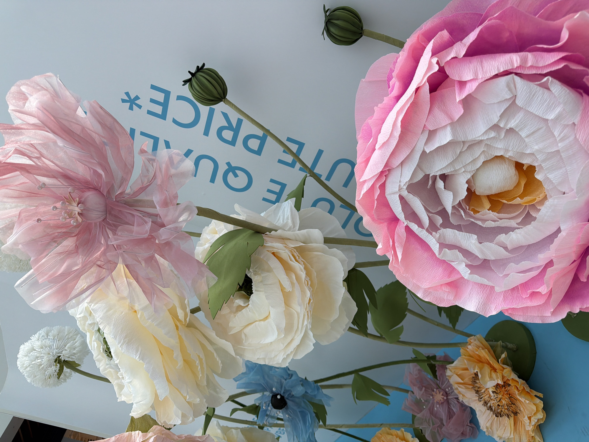 The image shows a close-up, low-angle view of an arrangement of large paper flowers against a light blue surface with partial lettering. The flowers are of various colors including pink, white, blue, and yellow, and include both open blossoms and closed buds. The lettering on the light blue surface includes the words "QUALITY PRICE." The scene appears to be indoors. While not explicitly stated in the image, the prompt implies a connection to Kommunarka, Russia, which is where the photo may have been taken.