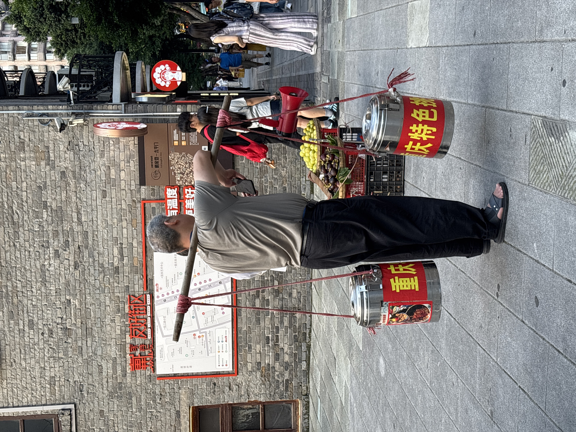 An adult male, with gray hair and a light-colored t-shirt and dark pants, is walking on a paved sidewalk, carrying a wooden pole across his shoulders. Two cylindrical, stainless steel containers are suspended from the ends of the pole by red ropes. Each container has a red label with yellow Chinese characters. The container on the left has an image of food on its label. The background features a brick wall with a red sign and a map, as well as other pedestrians and a building with a spiral staircase. The scene appears to be in a commercial area of Yuzhong District, China, indicated by the presence of signs and a map of the area.