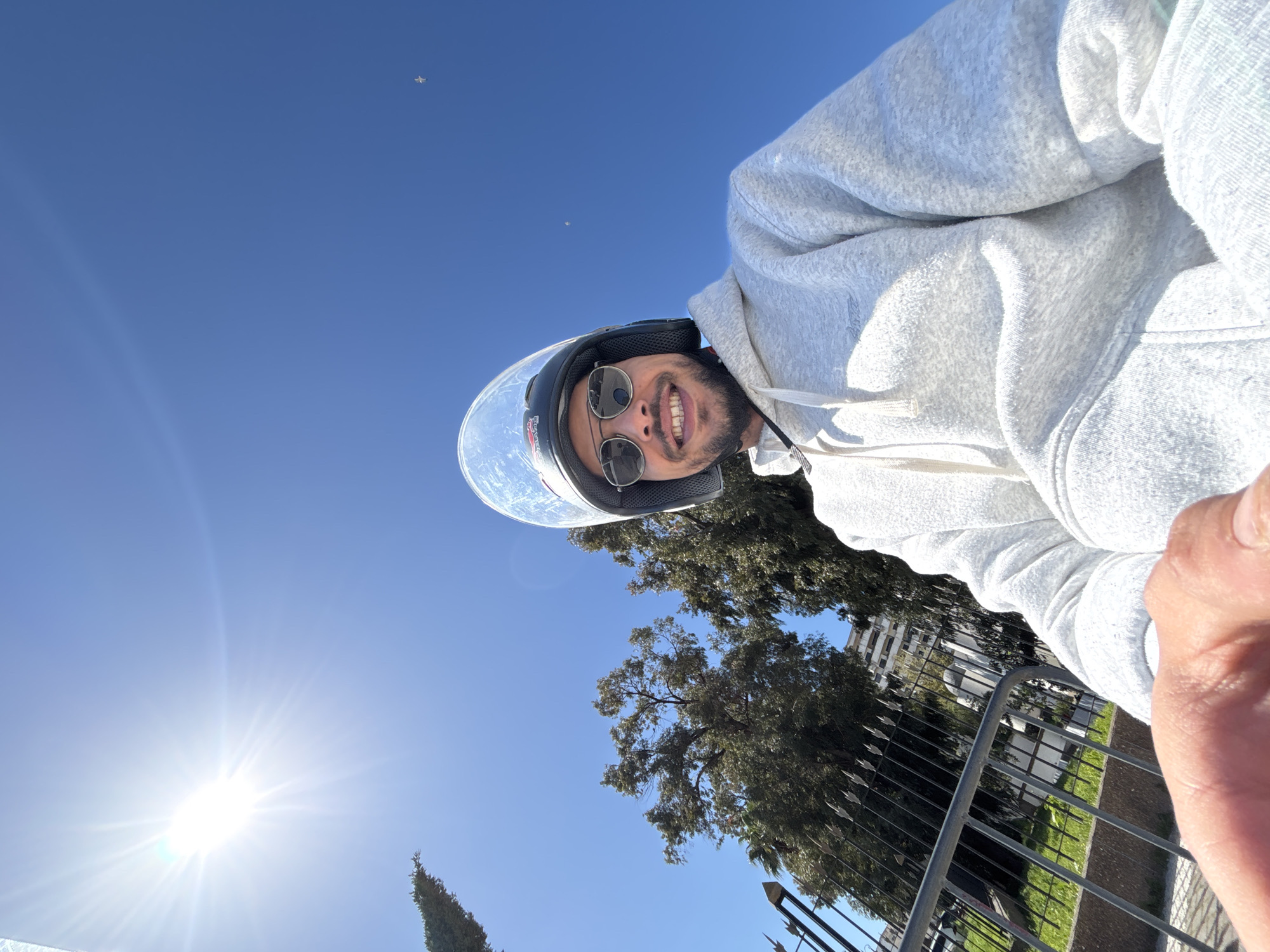 A male individual is captured in a close-up, low-angle selfie, positioned outdoors under a clear blue sky in the city of Tanger, Morocco. He is wearing a black helmet with a raised clear visor and dark, reflective sunglasses, covering his eyes. He has a dark beard and is smiling, showing his teeth. He wears a light grey hooded sweatshirt with drawstrings visible at the neck. His right hand is visible in the extreme lower foreground, suggesting he is holding the camera.
The environment is brightly lit by direct sunlight, which is prominently visible as a strong starburst in the upper left quadrant of the image. The sky is a deep, cloudless blue, with a faint, distant white object (possibly a bird or small aircraft) visible in the upper right. In the background, on the left, a tall, slender evergreen tree stands. Below it, a black metal fence with decorative spikes runs horizontally, separating the foreground from a green area. Beyond the fence, several large trees with dense green foliage are visible. A multi-story urban building, light in color, can be seen partially obscured by the trees and fence in the mid-distance. The setting appears to be an urban park or green space adjacent to a developed area within Tanger.