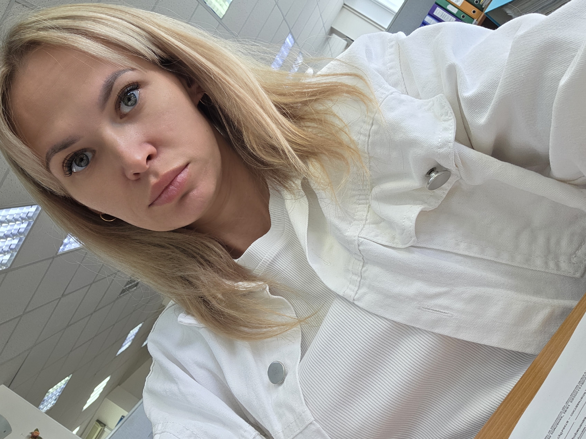 A young blonde woman is captured in a close-up, slightly angled selfie, set indoors, likely within an office environment in Chaikovsky, Russia. She has fair skin, light-colored eyes (appearing blue or grey), and prominent long eyelashes. Her gaze is directed towards the camera with a neutral, almost contemplative expression. She is wearing a white ribbed top beneath an open white jacket, which appears to be made of denim or a similar fabric, featuring silver-toned snaps or buttons. A small golden hoop earring is visible on her right ear.
The background shows a typical office ceiling with grid-patterned tiles and rectangular fluorescent light panels. In the upper right corner, a shelf holds several stacked binders or files in various colors (orange, green, purple, blue), further indicating a workspace. On the lower right, a wooden desk surface is visible with a piece of paper resting on it. Partially legible Cyrillic text can be seen on this paper, including acronyms like "ИНН" (Individual Taxpayer Number) and "ОГРН" (Main State Registration Number), which are common on official Russian documents. The overall lighting is bright and even, consistent with an artificially lit interior.