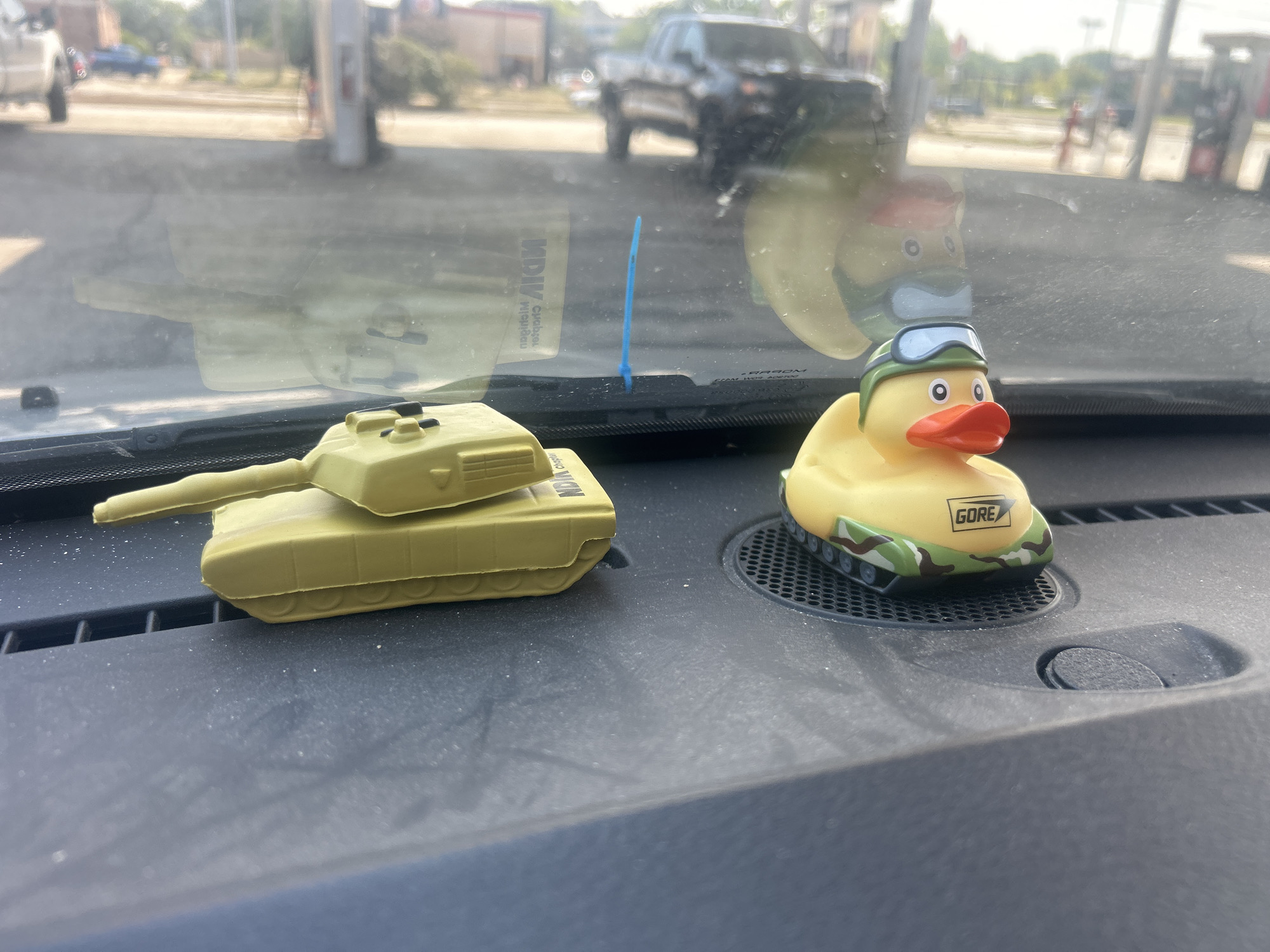 This is a close-up, eye-level shot of two toy military-themed objects on the dashboard of a vehicle, likely a car or truck, parked at a gas station in Willoughby, United States. The dashboard is made of dark, textured plastic and is covered in a fine layer of dust, with some white flecks scattered across it.
On the left is a tan-colored toy tank with a prominent barrel extending to the left. The tank is detailed with visible tracks.
To the right of the tank is a yellow rubber duck wearing a green helmet with blue goggles and an orange beak. The duck is perched on a grey circular grate. The base of the duck toy is camouflaged with green and brown patterns. A small white rectangle with black text, reading "GORE," is visible on the side of the duck's base.
In the background, through the windshield, a gas station is visible. There are gas pumps, a building, and several vehicles, including a dark-colored pickup truck, a white SUV, and a blue sedan. The lighting suggests it is daytime with bright sunlight. A thin, bright blue plastic zip tie stands upright on the dashboard in front of the windshield. The reflection of the outside environment is visible on the windshield, including what appears to be the silhouette of the tank and possibly the duck.