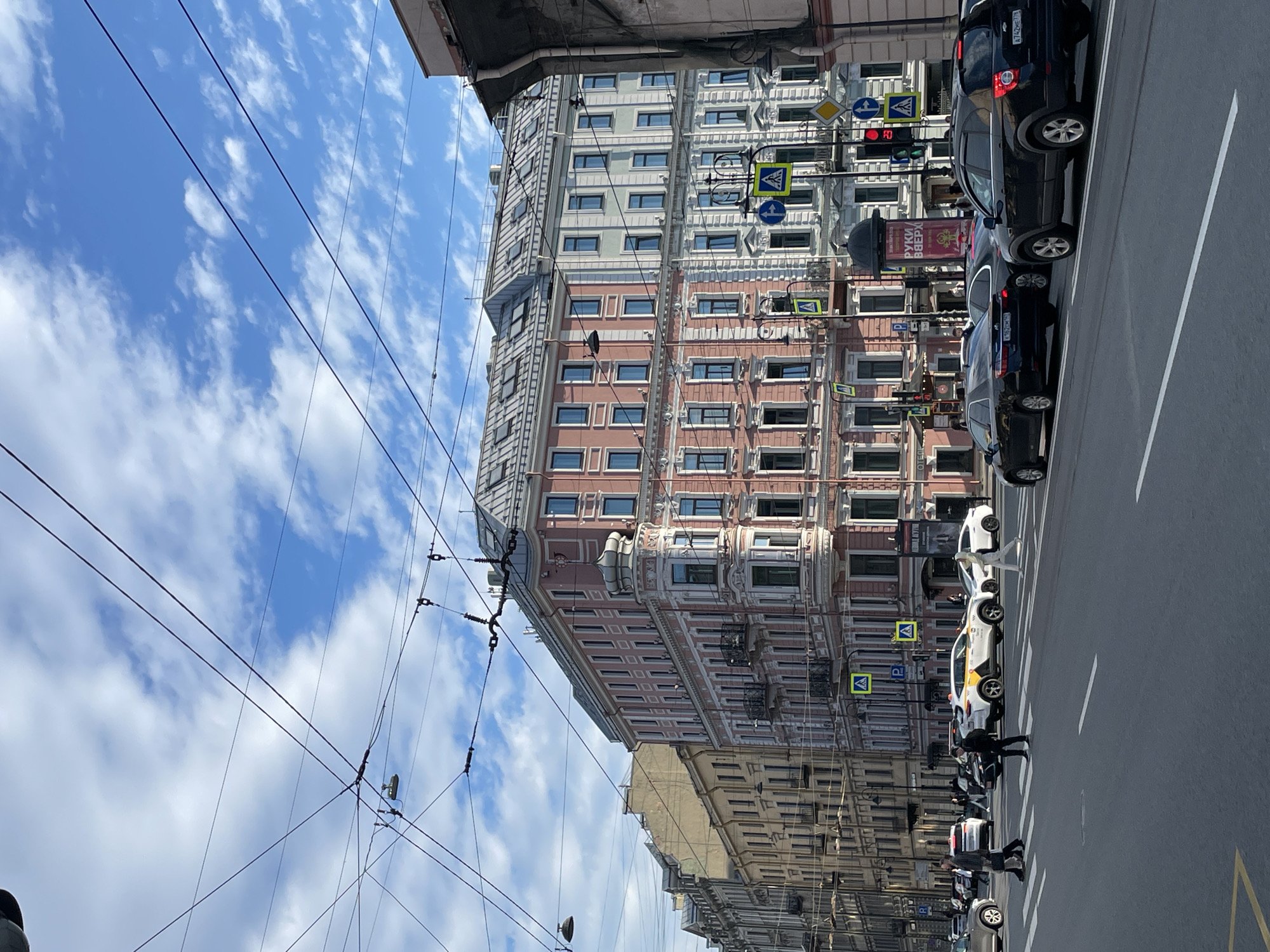 The image depicts a street scene in Saint Petersburg, Russia. The view is oriented vertically, showcasing tall, ornate buildings lining the street. The architecture includes details such as decorative window frames and varying colors, with one building featuring a predominantly pink and white facade. Above, a bright blue sky is filled with scattered clouds. Utility wires are visible stretching across the sky.
The street is paved and marked with lanes, with vehicles parked along the right side. Cars of varying colors including black and white are present. Pedestrians are seen crossing the street at a crosswalk marked by yellow pedestrian crossing signs. A traffic light is visible showing red. There are also street signs that indicate traffic regulations. A large billboard or sign is present which reads “РУКИ ВВЕРХ” in red lettering. The image is taken at ground level, with a slight upward angle towards the buildings.