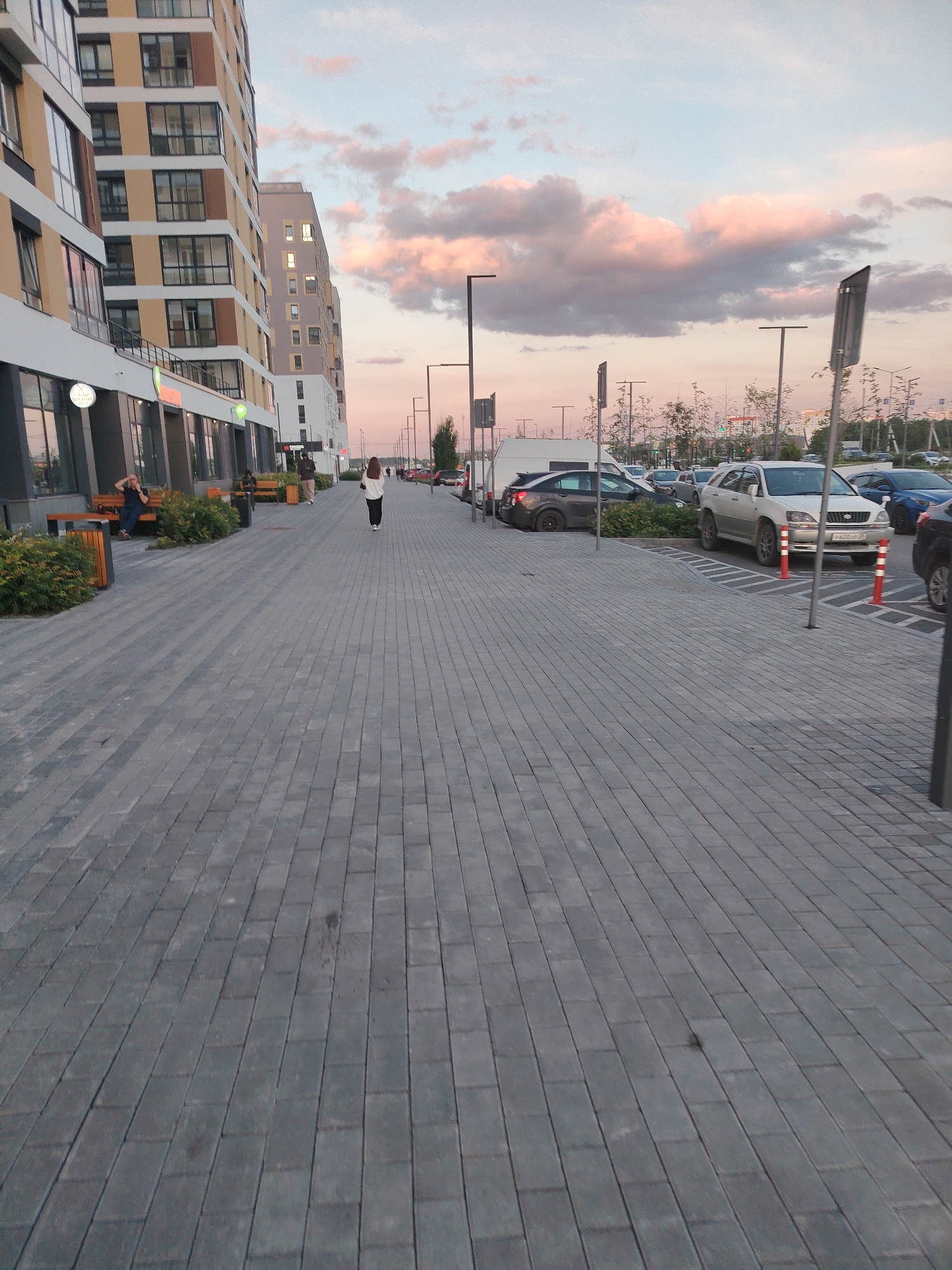 The image shows a wide, paved sidewalk with gray brick pavers in an urban setting. On the left, a modern apartment building with a beige and brown facade and large windows is visible. Several people are present: one person is sitting on a bench outside the building, another is standing near a trash receptacle, and a third person is walking away from the camera down the sidewalk. A fourth person, a woman with long brown hair, is walking towards the camera on the sidewalk.
To the right of the sidewalk, a parking lot is visible with several cars, including a silver SUV, a dark sedan, and a blue car. Orange traffic cones are placed in some of the parking spaces. Streetlights and trees line the parking lot. The sky above is a pale blue with soft pink and gray clouds, indicating it is either sunrise or sunset. The overall scene appears to be an outdoor public space in a residential or commercial area of a city.
