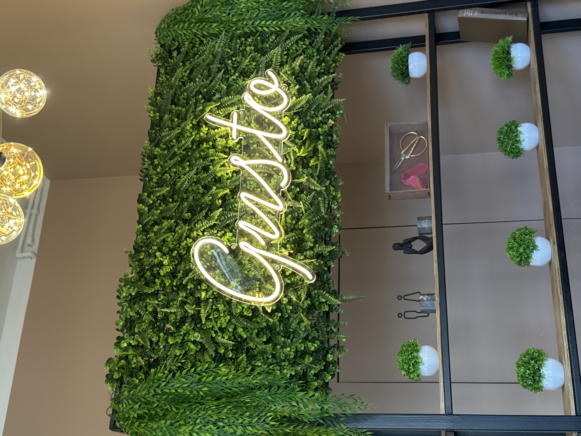 The image shows a neon sign that spells "Gusto" in cursive, glowing white, mounted on a wall covered in artificial greenery. The sign is positioned above two shelves of a dark metal shelving unit. The top shelf contains two small potted plants, a set of two stylized human figures, a decorative object of a person in a thoughtful pose, and a small wooden box with fabric and scissors inside. The second shelf holds one small potted plant. The third shelf has two small potted plants and a book. The background wall is a neutral beige color. The scene is illuminated by warm, ambient light, possibly from overhead fixtures visible at the top of the image. The location of Патрушева, Russia is not discernible from the image content.