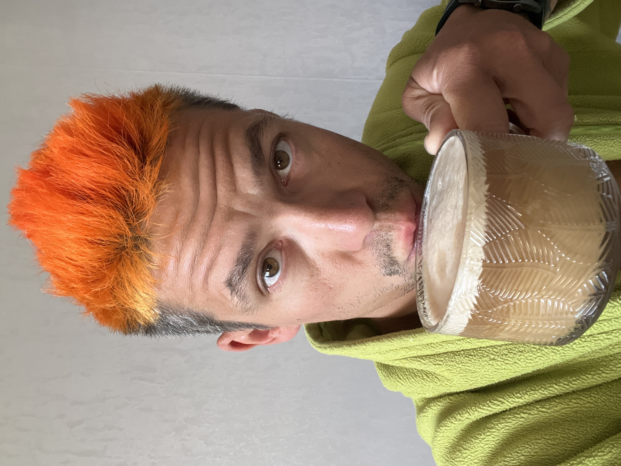 An indoor, eye-level shot captures a man with striking, bright orange hair and partially graying sides, looking directly at the camera. He wears a vibrant lime green fleece jacket. His brown eyes are wide open, and his forehead shows some wrinkles, suggesting a direct or slightly surprised expression. He has light stubble on his chin and jawline.
He is holding a clear, textured glass mug filled with a frothy, light brown beverage, likely coffee or a similar drink. The mug is raised to his mouth, suggesting he is either about to take a sip or has just finished one. A watch is visible on his left wrist. The background is a plain, light-colored wall, indicating an indoor setting. The lighting is even, illuminating his face and the details of his clothing and the mug. The scene takes place in Chaikovsky, Russia.