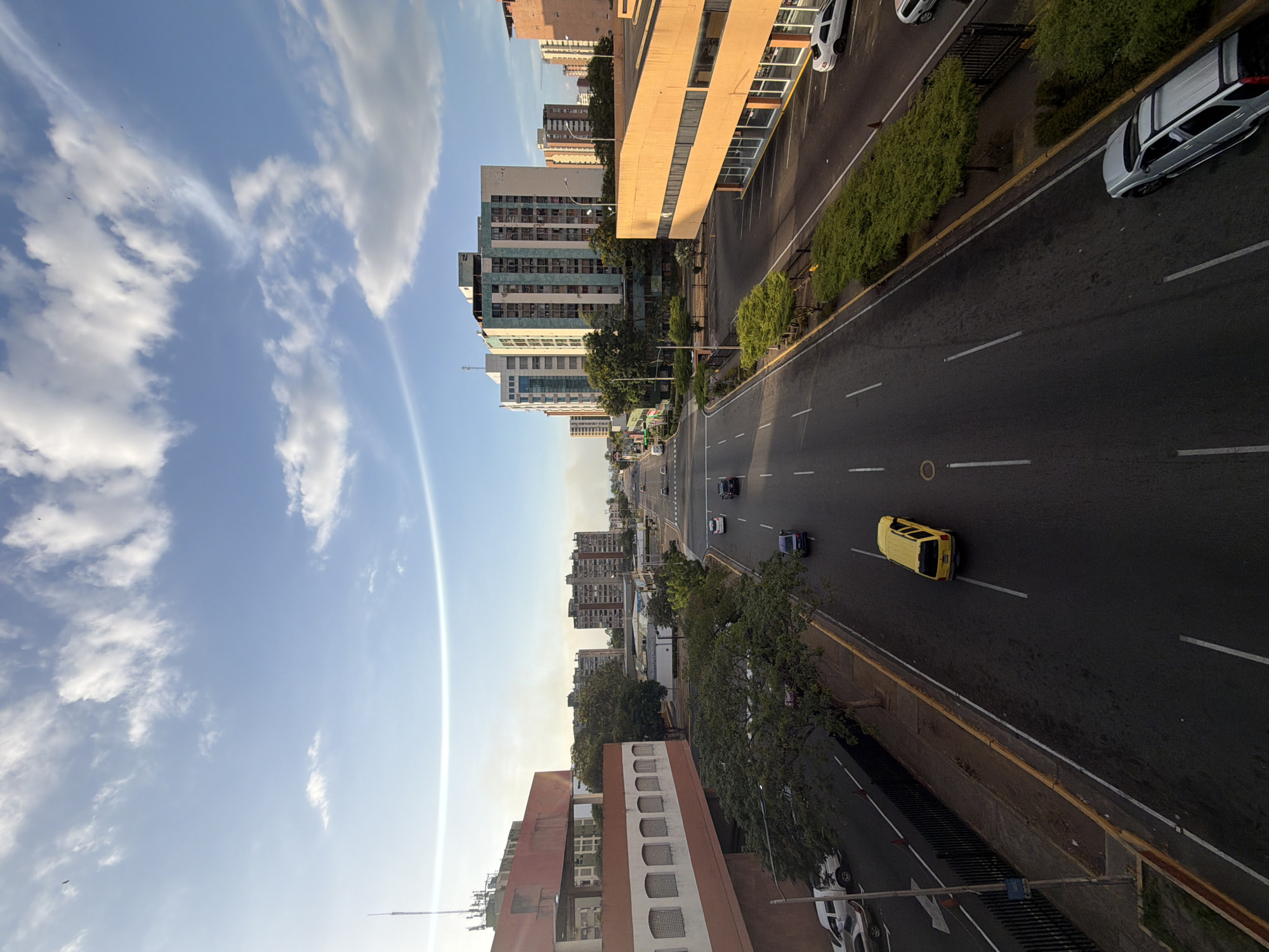 The image shows an elevated view of a street in Ciudad Guayana, Venezuela, with several buildings visible along both sides. The sky is visible at the left of the image, it is blue with white clouds and contrails. A multi-lane road runs through the center, with several vehicles visible including a yellow car in the lower half, also a black and a blue car on the upper half of the road. Buildings of varying sizes and architectural styles are arranged along the sides of the road, with some trees and greenery interspersed. The overall impression is that of a typical urban scene on a clear day.