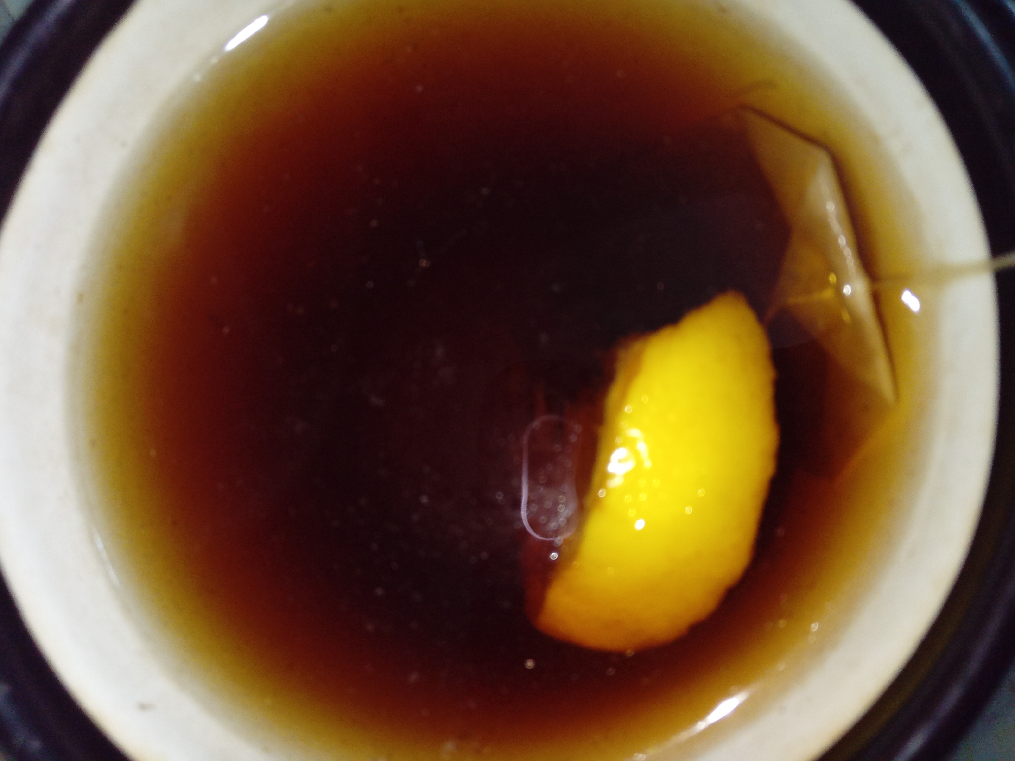 The image shows a close-up overhead view of a mug of dark tea. A slice of bright yellow lemon floats on the surface of the tea, positioned to the left of the center of the mug. Partially submerged in the tea near the lemon slice is a tea bag, its paper tag folded and visible just below the liquid's surface. The mug itself is light-colored with a beige rim, and it sits within a darker outer container, possibly a coaster or another mug. The lighting casts reflections on the surface of the tea, creating bright, circular highlights. The scene is static and appears to be indoors, with no discernible elements that would place it specifically in Kitskany, Moldova.