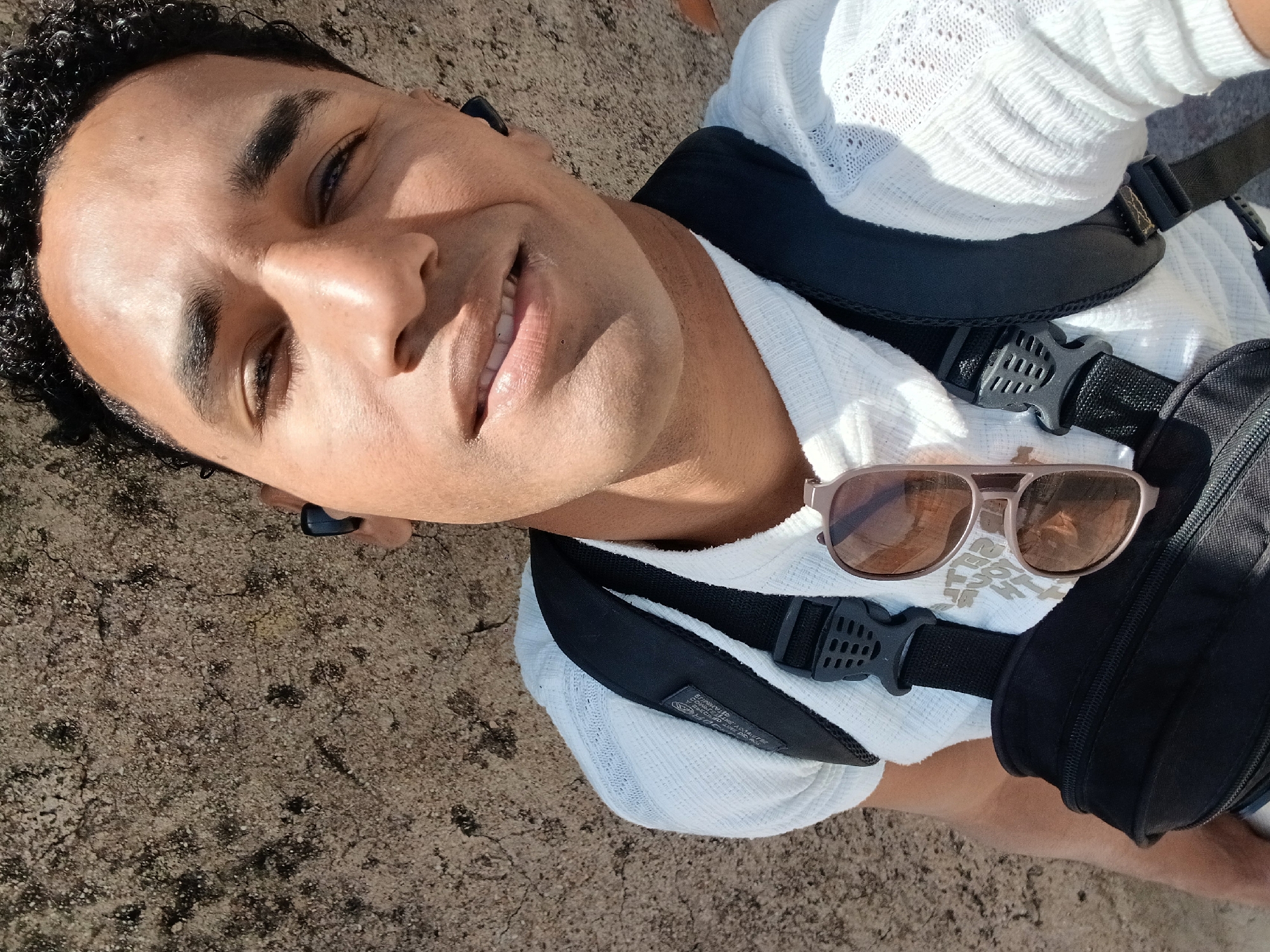 A selfie of a man with dark curly hair and brown skin. He is wearing a white open-weave sweater and a black backpack. Sunglasses rest on one of the backpack straps. He has wireless earbuds in his ears. The background appears to be a concrete or stone surface. The location is potentially Porlamar, Venezuela. No specific landmarks are visible to confirm this.