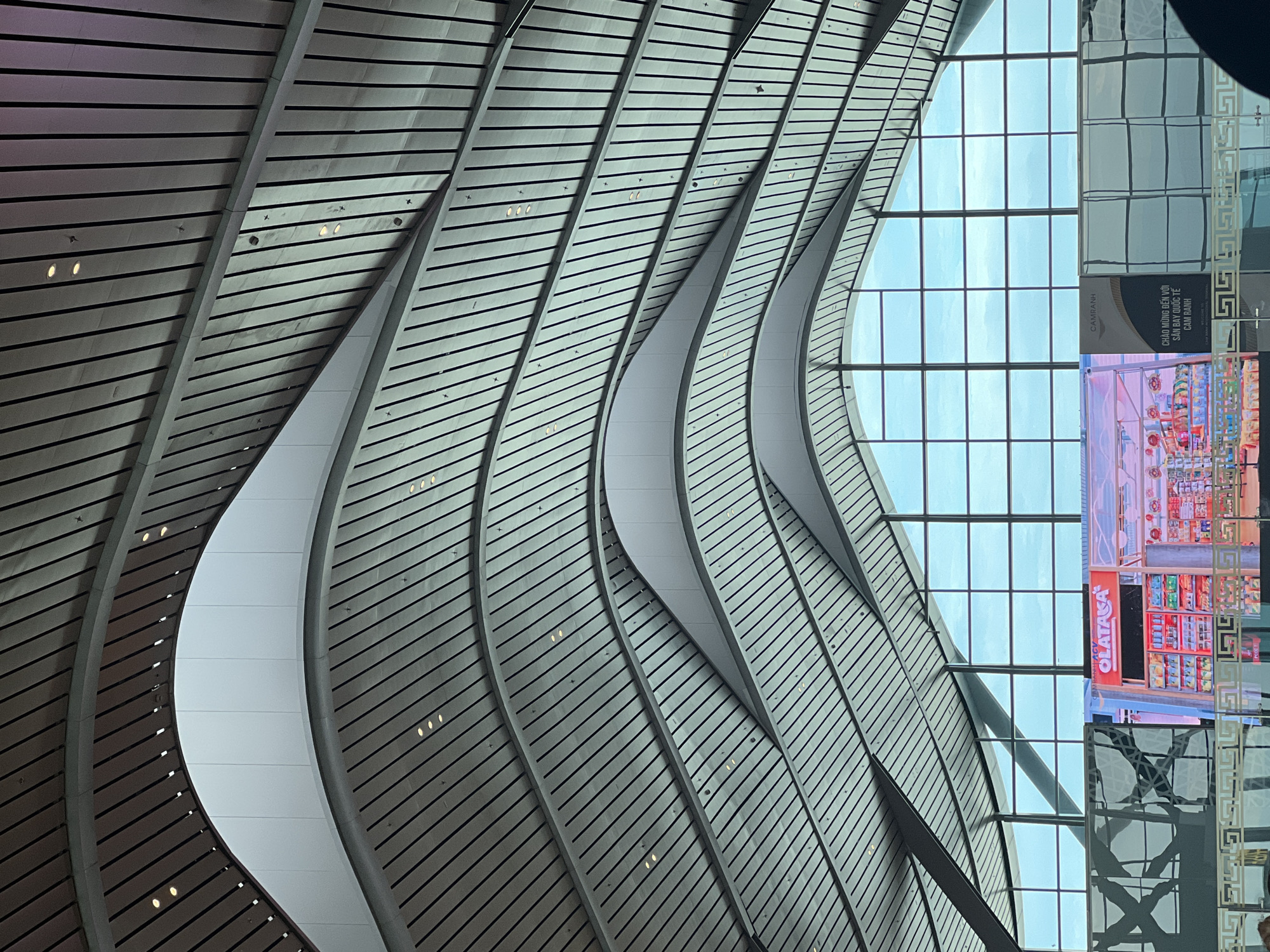 The image displays the interior of a modern architectural space, likely an airport terminal, characterized by a striking, curvilinear ceiling or upper wall. The dominant feature is a series of undulating panels, light gray or white in color, arranged horizontally with dark stripes, creating a slatted appearance. These panels form graceful, wave-like curves that extend across the frame, with small, circular lights recessed within them.
To the right, a large section of grid-patterned glass windows offers a view of a bright, clear sky, indicating daytime. Below these windows, reflections and structural elements are visible, along with a large digital display screen. The screen shows the interior of a retail store, possibly a pharmacy, featuring various products and the prominent text "OLATASA" in red letters, with "PHARMACY" above it.
A banner or sign positioned near the windows prominently displays the Vietnamese text "CHÀO MỪNG ĐẾN VỚI SÂN BAY QUỐC TẾ CAM RANH," which translates to "Welcome to Cam Ranh International Airport." The words "CAM RANH" and "CAMRANH" are also visible on this sign. Decorative golden patterns adorn some of the architectural elements adjacent to the windows. The overall scene conveys a modern, spacious, and well-lit environment, consistent with a contemporary international airport in Bac Cam Ranh Ward, Vietnam. No people are visible in the image.