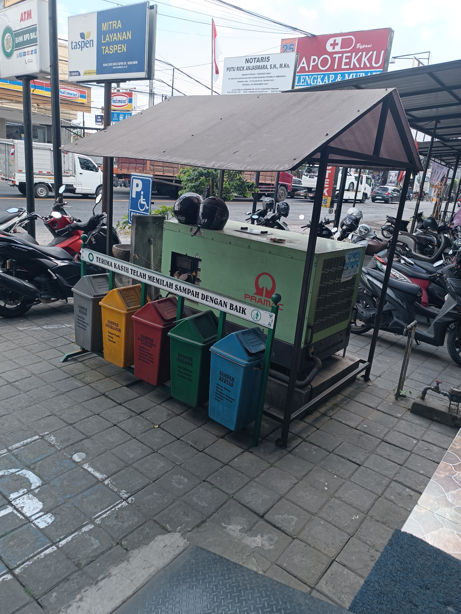 A generator unit sits on a metal platform in front of a row of five colored trash bins. The bins are labeled for different types of waste, including "SAMPAH RESIDU," "SAMPAH ANORGANIK," and "SAMPAH ORGANIK." The generator is green and has the brand name "PRAMAC" visible. A horizontal sign above the bins reads "TERIMA KASIH TELAH MEMILAH SAMPAH DENGAN BAIK" (Thank you for sorting your trash properly). Two black helmets rest on top of the generator. The scene appears to be outdoors, with a paved ground covered in square concrete tiles. Behind the generator and bins, several motorcycles are parked under a covered structure. In the background, there are buildings and a street with traffic. The overall setting suggests a public area, possibly near a market or commercial district in Tabanan, Indonesia, where waste sorting is encouraged.
