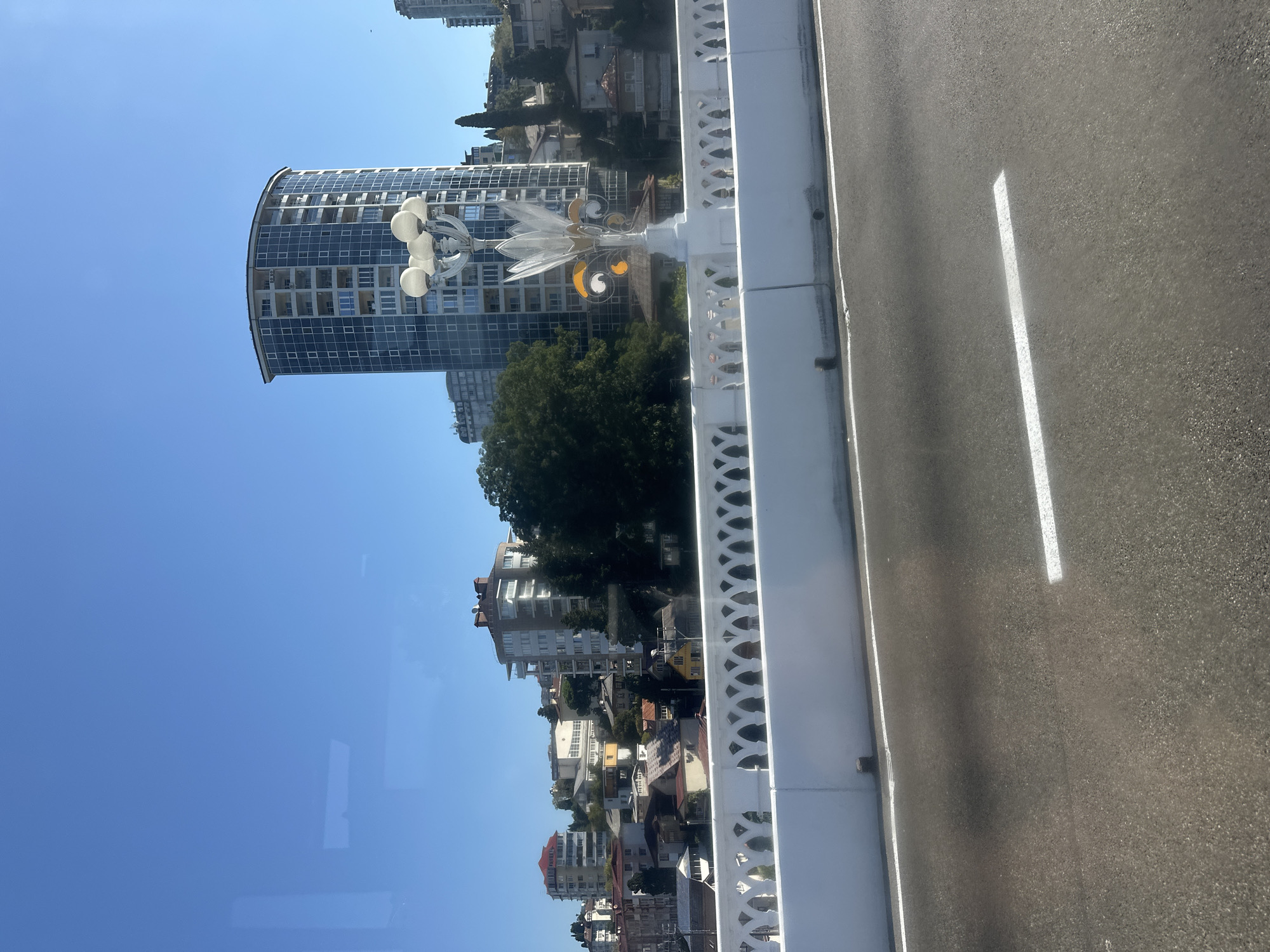 A view of the Khosta Bridge in Sochi, Russia, shows a white decorative balustrade on the bridge, with a multi-headed street lamp featuring glass accents. Beyond the bridge, a cityscape unfolds with various buildings, including a modern, curved, glass-fronted high-rise, other apartment buildings, and smaller houses scattered across a hillside. The sky is clear and blue. The foreground shows a section of asphalt road with a white lane marking.