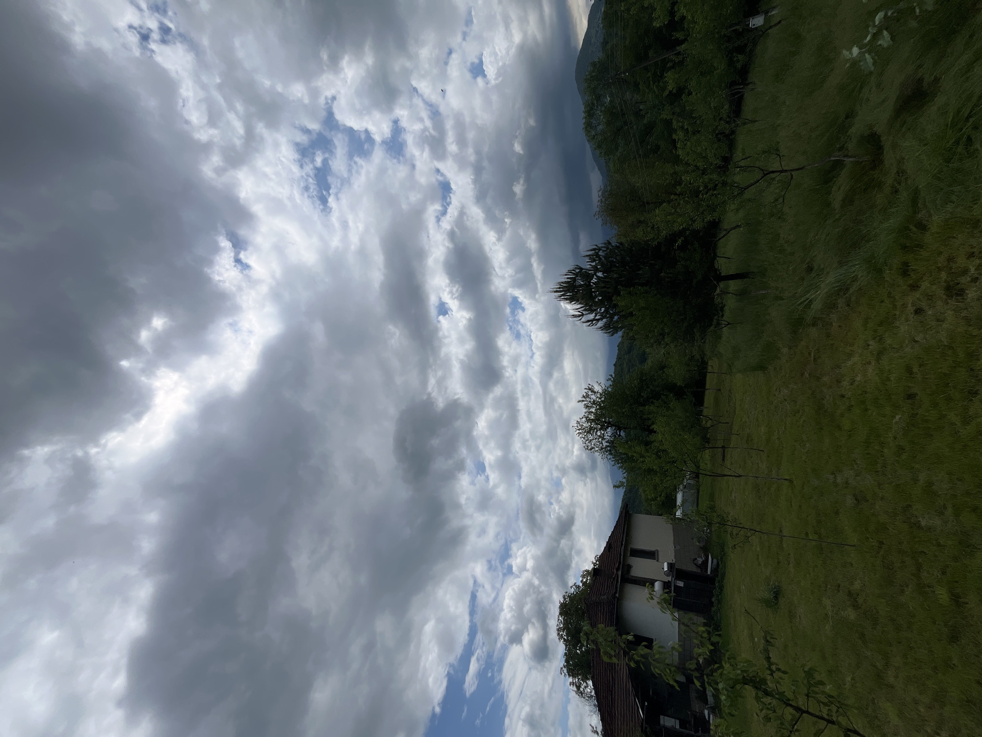 The image presents a view of a landscape in Shopite, Bulgaria. The foreground consists of a green meadow with tall grass and several young trees. In the middle ground, there is a two-story house with a brown roof. The house is surrounded by trees and some kind of fence. The background is dominated by a dramatic sky filled with grey clouds, but with patches of blue visible. The sky occupies a significant portion of the image, suggesting stormy weather. The image is taken from a slightly elevated position, looking down onto the house and meadow. There is a mountain ridge, covered by trees, in the distance.