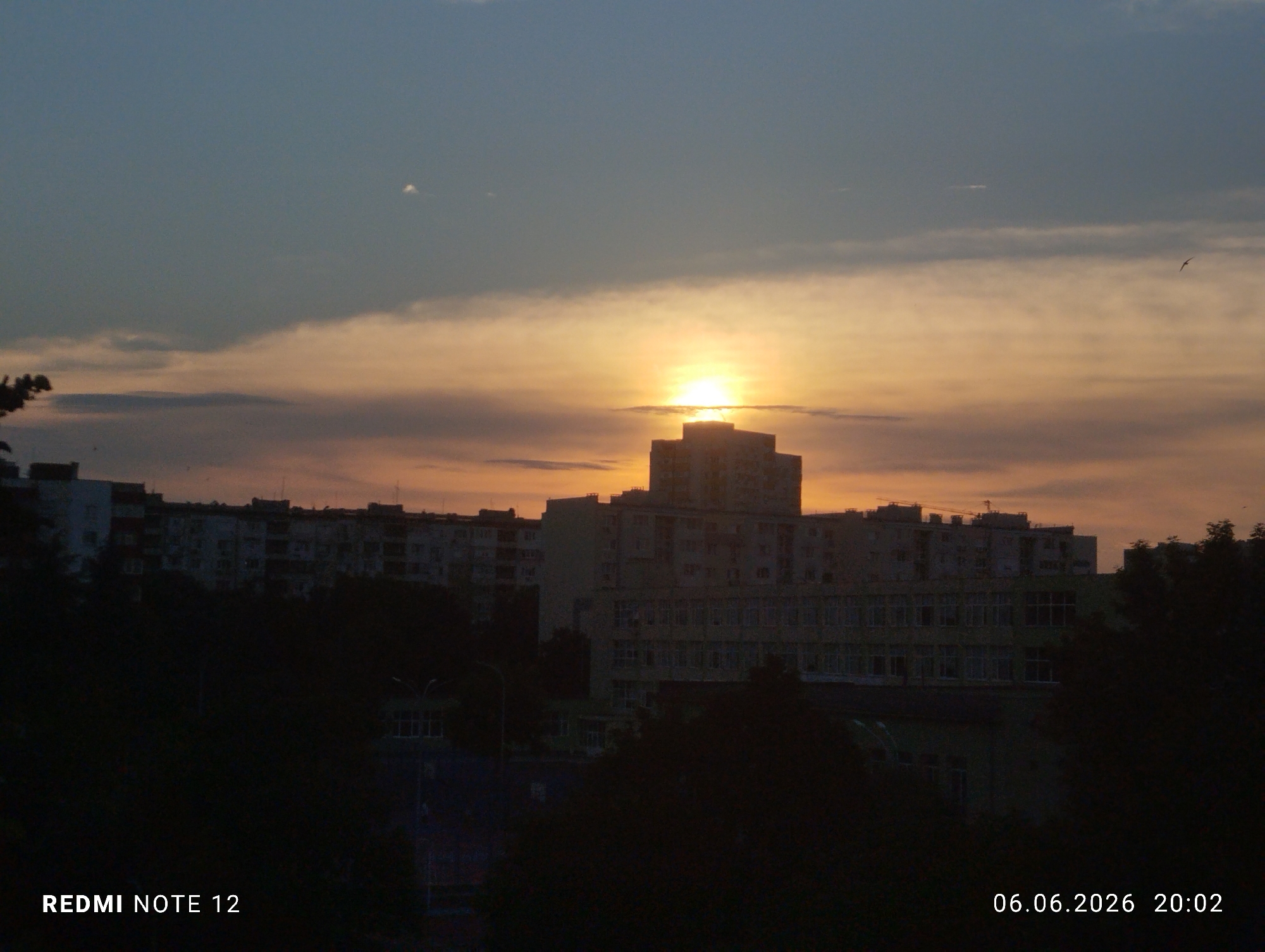 The image captures an urban sunset scene in Burgas, Bulgaria, at 20:02 on June 6, 2026, taken with a Redmi Note 12 device. The sky displays a gradient from dark blue-grey at the top to vivid orange, yellow, and peach tones near the horizon. The setting sun, a bright yellow disc, is partially visible above a prominent, multi-story building, casting it and other structures into silhouette.
A large, multi-story building with a stepped or tiered roofline is centrally located, directly in front of the sun. To its left and right, several other multi-story residential or commercial buildings are visible, heavily shadowed. One building in the mid-ground on the right has discernible window patterns, and a construction crane is faintly visible atop a more distant structure. The lower third of the image consists of dense, dark silhouettes of trees and foliage, obscuring the ground level and lower portions of the buildings. A street light with an ornate top is partially visible as a silhouette amidst the foreground trees in the lower left. Faint clouds streak across the sky, and a small, dark bird is visible in flight in the upper right portion of the illuminated sky. A small, indistinct white object or contrail is present in the upper sky. No individuals or specific actions are discernible.