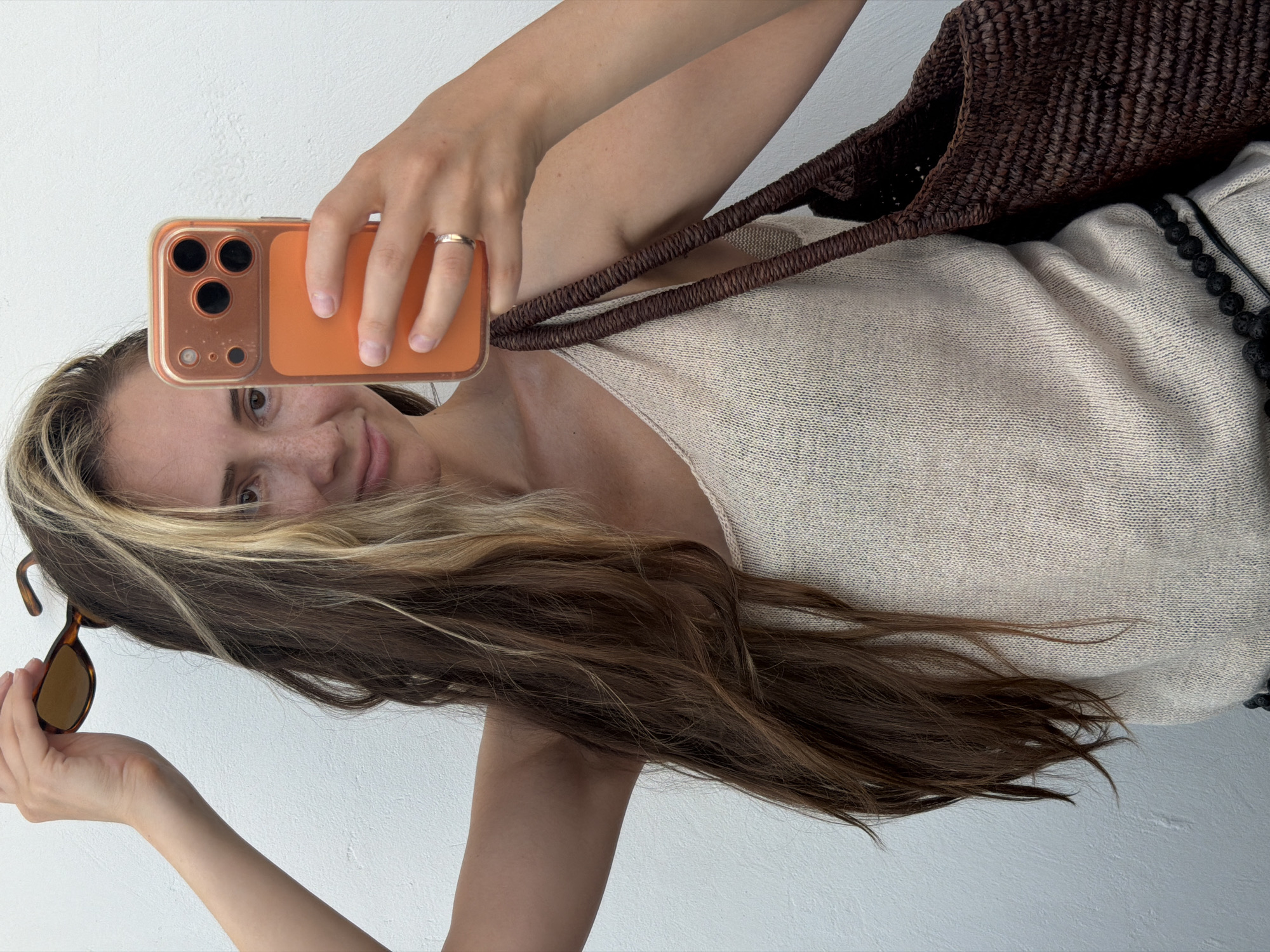 A young woman is taking a selfie in front of a white wall. She is holding a smartphone with an orange case in her right hand. Her left hand is resting on top of her head, holding sunglasses. She is wearing a light-colored knitted tank top and a woven brown bag is slung over her left shoulder. She has long, multi-colored brown hair. Her left arm is bent and raised. A ring is visible on her right ring finger. The background is a plain white wall.