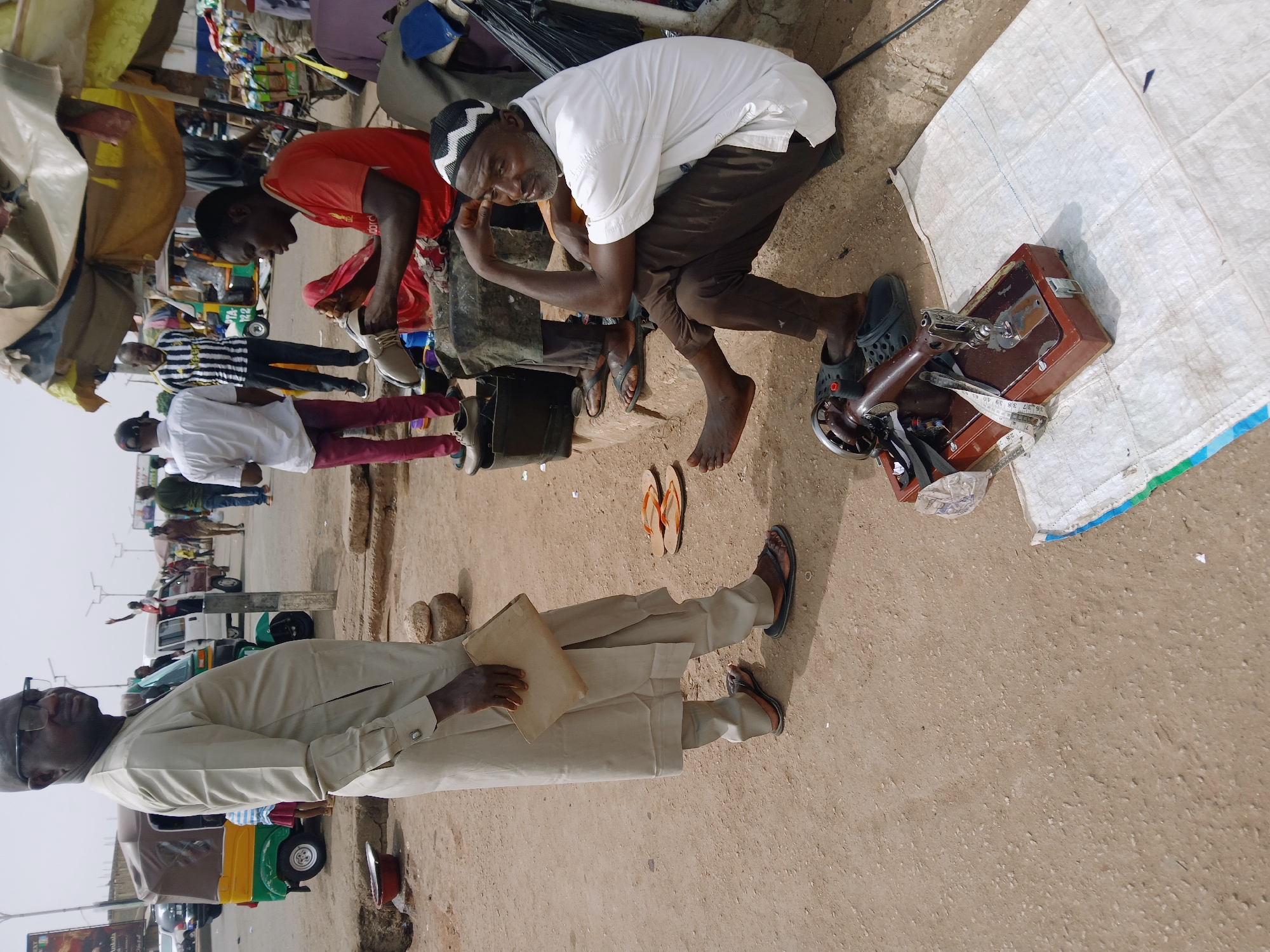 A street scene in Jos, Nigeria, shows a man in a cream-colored traditional outfit holding a document. He stands on a dusty ground near a road. In the background, there are auto rickshaws and pedestrians. Near him, another man kneels next to a sewing machine placed on a white sack. A second man is seated close by, appearing to be fixing shoes. Further in the background, pedestrians walk alongside vehicles. The overall environment suggests a typical roadside setup for small-scale commerce and services.