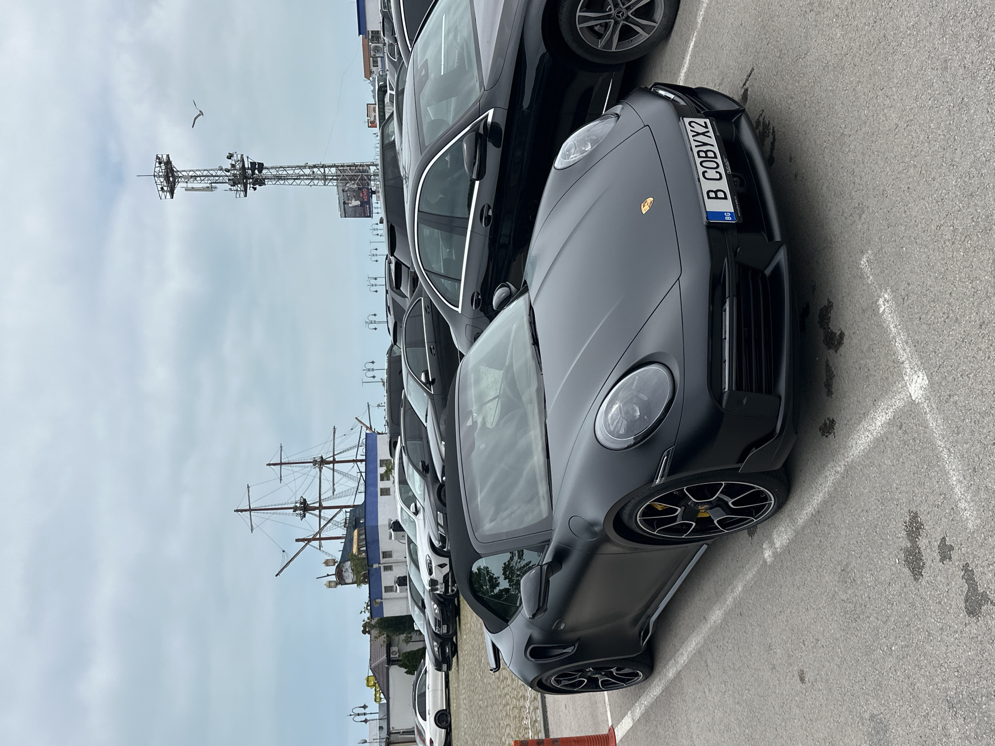 The image displays a line of parked cars in an outdoor asphalt lot, likely in Varna, Bulgaria, under an overcast sky. In the foreground, a sleek, matte black Porsche 911 convertible, possibly a Targa or Cabriolet model, is prominently featured. It has striking yellow brake calipers and multi-spoke black and silver wheels. The Porsche's front license plate is clearly visible, reading "B C0BYX2" with the "BG" country code, indicating Bulgarian registration.
Behind the Porsche, several other vehicles are parked in a row, predominantly dark-colored sedans and SUVs, including what appears to be a black Mercedes-Benz sedan.
In the mid-ground, to the left, a tall metal communication or observation tower stands against the grey sky, equipped with various antennas and platforms. A single bird, possibly a seagull, is captured flying in the sky near this tower. To the right and further in the background, the intricate masts and rigging of a large sailing ship structure are visible, suggesting a replica or a themed attraction, which aligns with a coastal setting.
The ground is an asphalt surface marked with white parking lines, showing some dark stains. The time of day is daytime, with consistently overcast weather. No people or active events are visible in the scene. A partial text "NEM" is faintly discernible on a billboard attached to the tower.