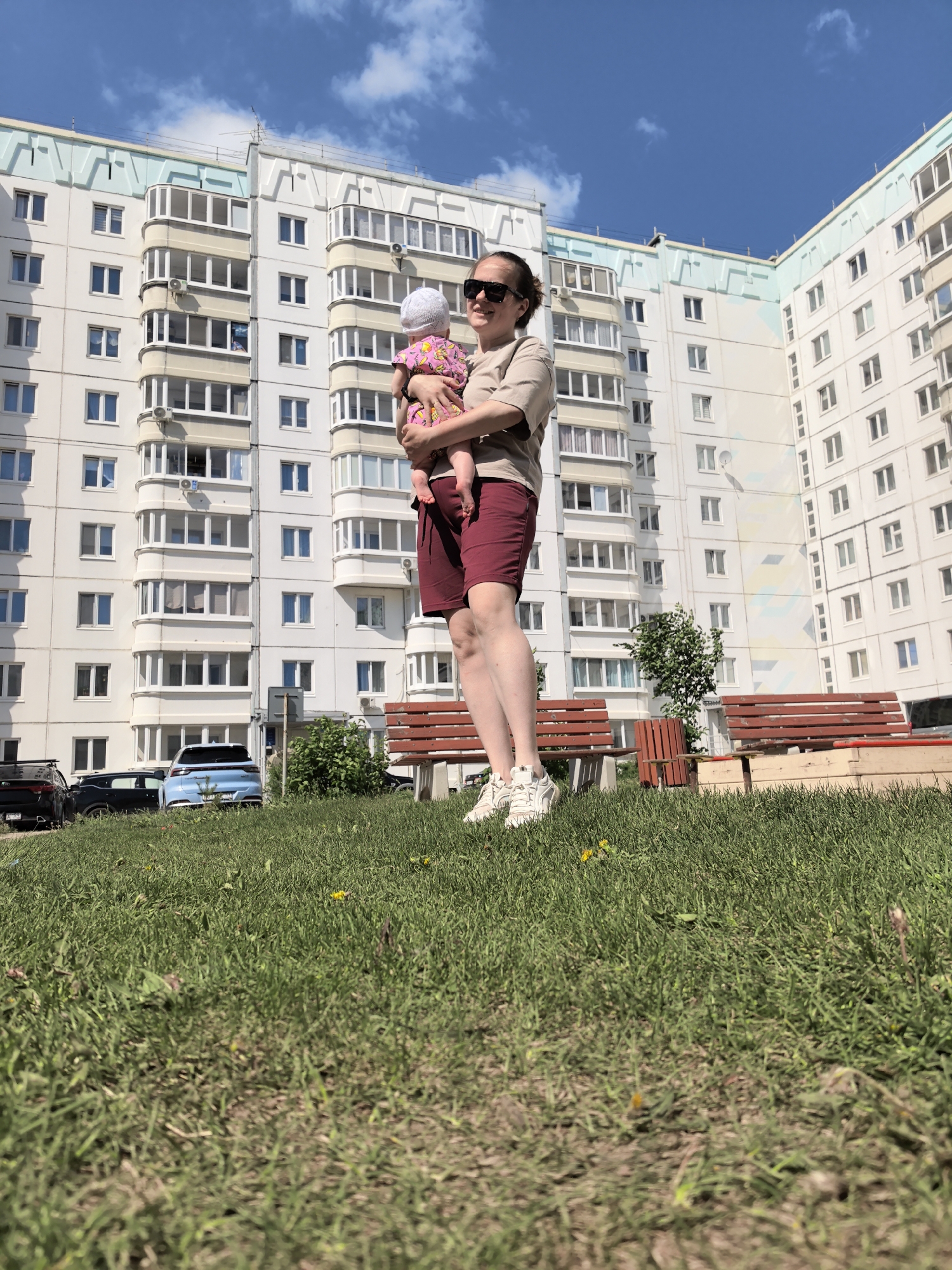 The image depicts a woman holding a baby, standing on a grassy lawn. In the background is a white apartment building with blue-green accents. Behind the woman are park benches and parked cars. The image appears to have been taken in the city of Фролы, Russia. The woman is wearing a beige shirt, maroon shorts, and white sneakers. She also wears sunglasses and has her hair pulled back. The baby is wearing a white hat and a pink floral outfit. They are standing outside in an open area during the day.