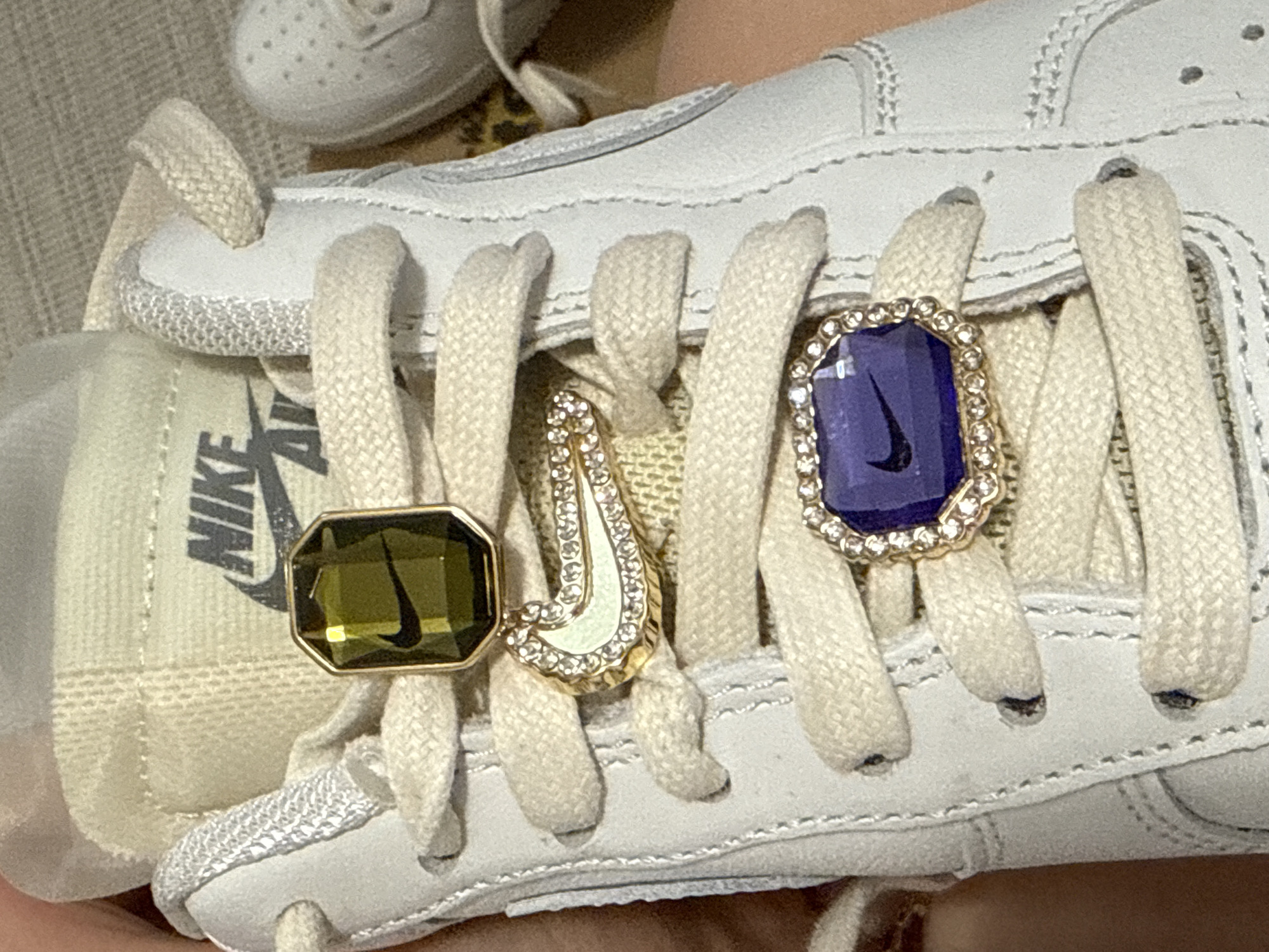 This image depicts a close-up view of a white Nike Air Force 1 shoe. The laces are also white and adorned with three decorative charms. The charm on the left is a dark green octagon with a gold border and a Nike swoosh design. The charm in the center is a curved Nike swoosh shape with a gold border and inlaid with small stones or rhinestones. The charm on the right is a blue octagon with a gold border featuring small stones or rhinestones around its edge, also containing a Nike swoosh design. The surrounding environment is out of focus, suggesting a domestic setting. The location of Hainburg an der Donau, Austria, is not discernible from the image.
