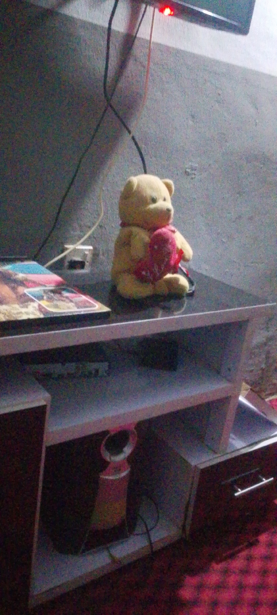 A fluffy yellow teddy bear with a red heart is sitting on a black glass-top TV stand in an indoor setting. The bear is positioned to the right of a stack of colorful books or magazines. The TV stand has multiple shelves, with one shelf holding a cylindrical object with a pink accent and a black cord. In the background, a textured grey wall is visible, with electrical wires running along it. A red LED light is illuminated on a black object at the top of the frame, possibly a television. The floor is covered with a red carpet with a diamond pattern. The scene appears to be a room in a home or similar dwelling.