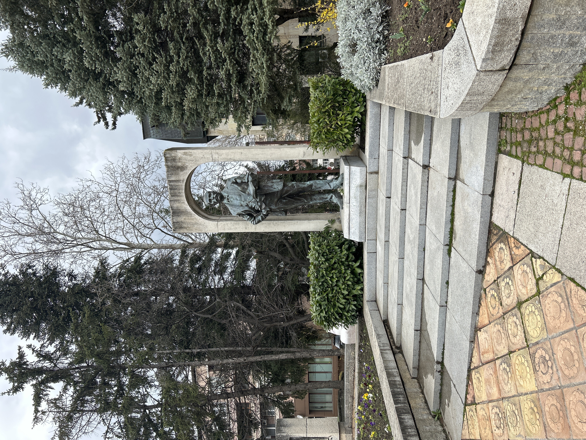 The image depicts a bronze statue of a man, likely a historical figure, standing within a concrete arch. The statue is located in a park or memorial area in the city of Shumen, Bulgaria. The statue is positioned at the top of a set of stone steps, with greenery and flowers planted on either side. Tall evergreen trees surround the monument, and other buildings are visible in the background. The steps lead to a tiled walkway. The sky is overcast.