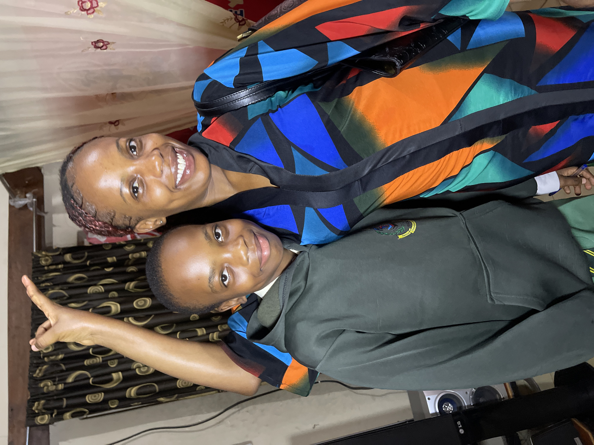 In the city of Jos, Nigeria, a young boy and a woman pose for a photo in an indoor setting. The boy, positioned on the left, wears a dark green hooded sweatshirt with a school crest on the left chest. The woman, on the right, is dressed in a vibrant, patterned robe featuring geometric shapes in shades of blue, orange, green, and red, with black accents. Her right arm is raised, with her hand visible above the boy's head. Behind them, curtains with a brown and gold pattern hang on the left, and a lighter, sheer curtain with red floral embroidery is visible on the right. To the left of the boy, a black LG speaker is partially visible. The lighting suggests an indoor environment, possibly a home or room.