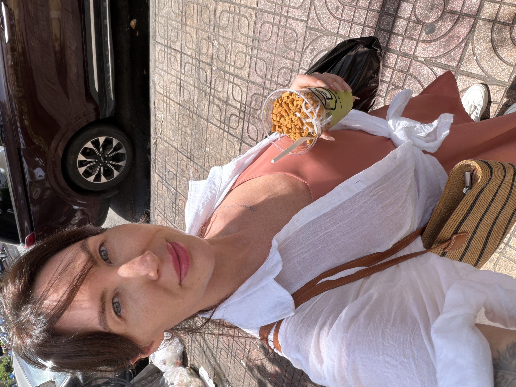In Nha Trang Ward, Vietnam, a woman of Caucasian ethnicity is captured in a high-angle, full-body selfie. She has fair skin with freckles, light blue eyes, and brown hair pulled back from her face, with some strands falling forward. She is wearing a coral-pink sleeveless dress, cinched at the waist, and an unbuttoned white linen shirt over it, tied at the front. A brown woven straw bag with leather straps hangs from her left shoulder, and a black plastic bag is held in her right hand. She is holding a clear plastic cup filled with a green beverage topped with crushed cookies and possibly coconut flakes, with a wooden stirrer. Her left hand has visible tattoos on the forearm. The background reveals a sidewalk paved with patterned tiles and a dark red vehicle, possibly a minivan, parked on the street. In the upper left corner, some white plastic bags are piled on the ground. The scene appears to be outdoors on a sunny day.