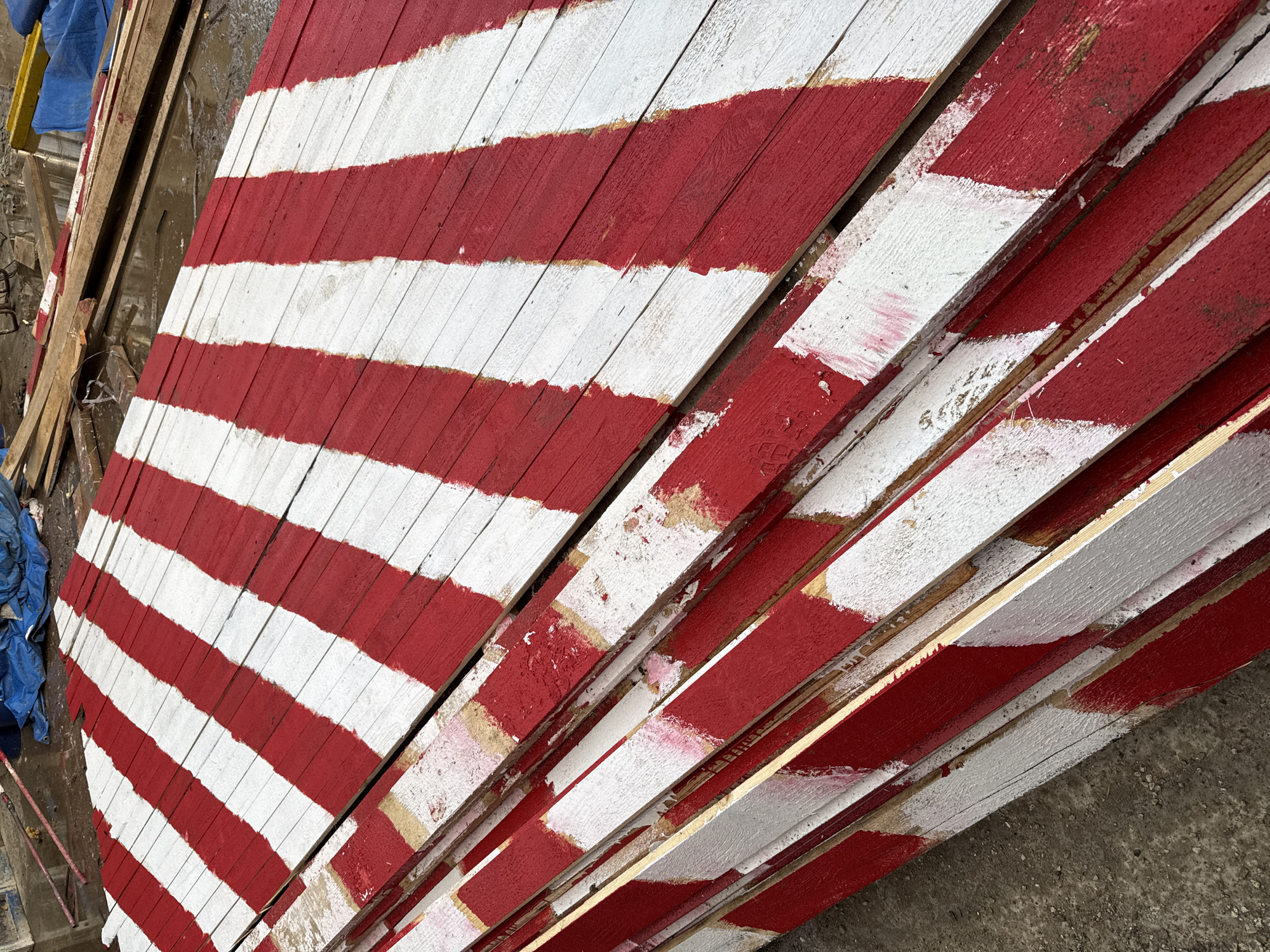 A stack of wooden planks, painted with alternating red and white diagonal stripes, lies on a wet, gravelly surface. The planks are rough and weathered, with visible wood grain and some areas where the paint has chipped away. In the background, other wooden materials, a blue tarp, and puddles of water suggest an outdoor construction or storage area. The scene is devoid of any people or animals. The location is not specified in the image.