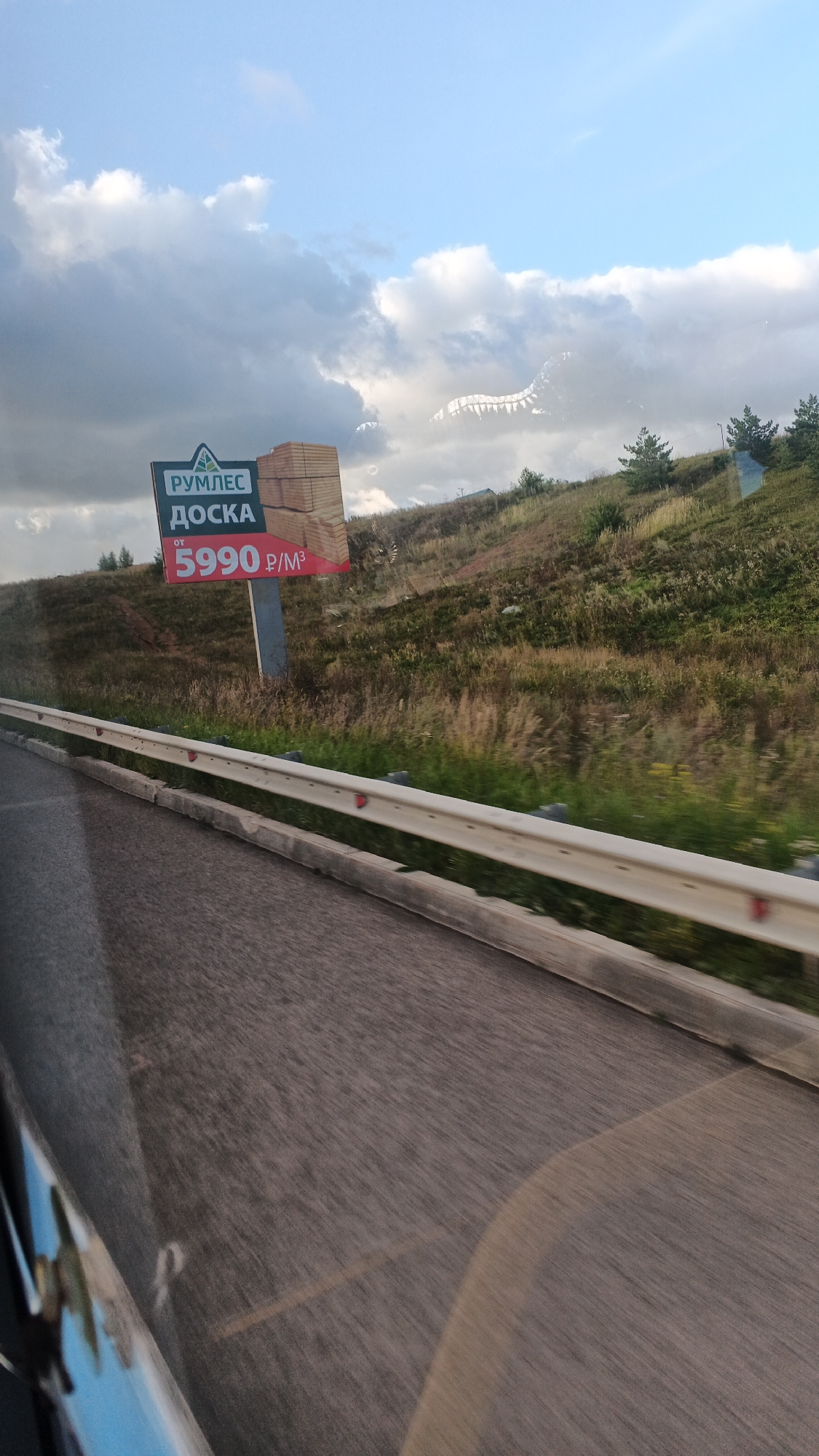The image captures a roadside billboard in Kamyennoye, Russia, advertising lumber. The billboard features the logo "РУМЛЕС", followed by "ДОСКА" (meaning "board"), and the price "ОТ 5990 Р/М³". To the right of the text, there are stacked wooden boards. The billboard is positioned on a grassy, sloped hillside, with scattered trees and brush. A metal guardrail separates the road from the hillside. The road itself is paved and appears to be a highway, with motion blur indicating the perspective is from a moving vehicle. The sky above is partly cloudy with a mix of blue sky and white clouds. The overall scene is set in a rural or semi-rural environment.