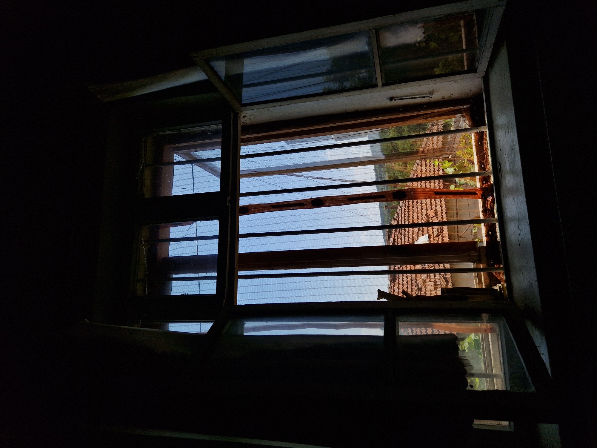 The image depicts an interior view looking out of a window. The window frame is old and appears weathered, with some damage visible on the right-hand side. The window is partially open, and through the bars, a rural landscape can be seen. The landscape includes a building with a tiled roof, trees, and a clear blue sky. The interior of the room is dark, with a heavy curtain visible on the left side, obscuring a large portion of the view. The overall impression is one of abandonment or neglect. The image was captured in Raduvene, Bulgaria, a town known for its rural setting and traditional architecture.