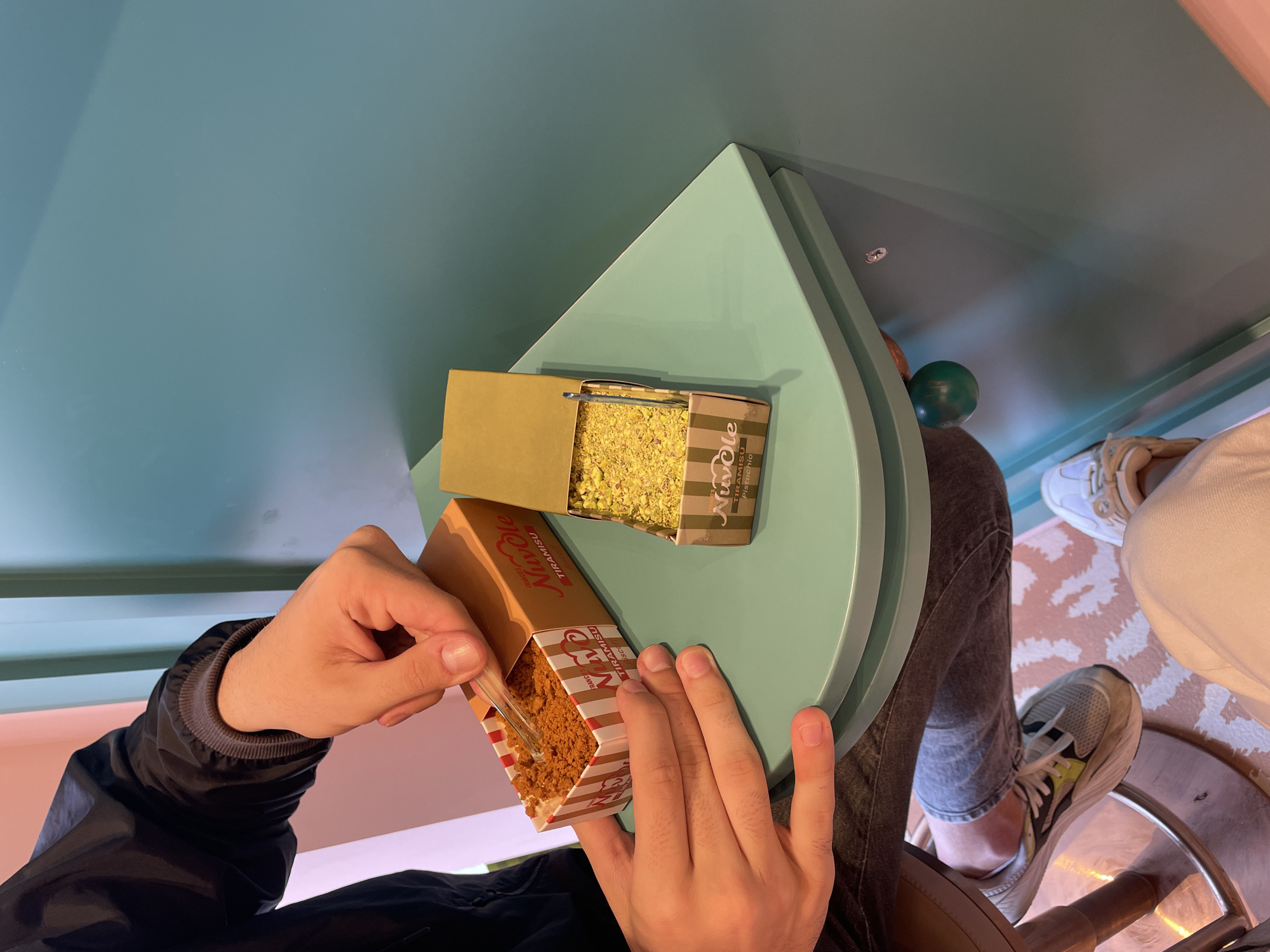 From a high angle, a person is seen eating a Tiramisu dessert in what appears to be a café or restaurant setting. The person, wearing a black jacket and grey jeans, is sitting on a stool with grey sneakers visible. The dessert, packaged in a brown and white striped box from 'Nuvole Tiramisu', is held by the person, who uses a small plastic fork to eat it. A box of pistachio tiramisu from the same brand is nearby. The table is a light green, kidney-shaped surface. Below the table, patterned carpet can be seen along with partial views of shoes of a second person. The background includes a light blue wall and an adjacent pink wall. The location is a Nuvole Tiramisu Shop in Sofia, Bulgaria.