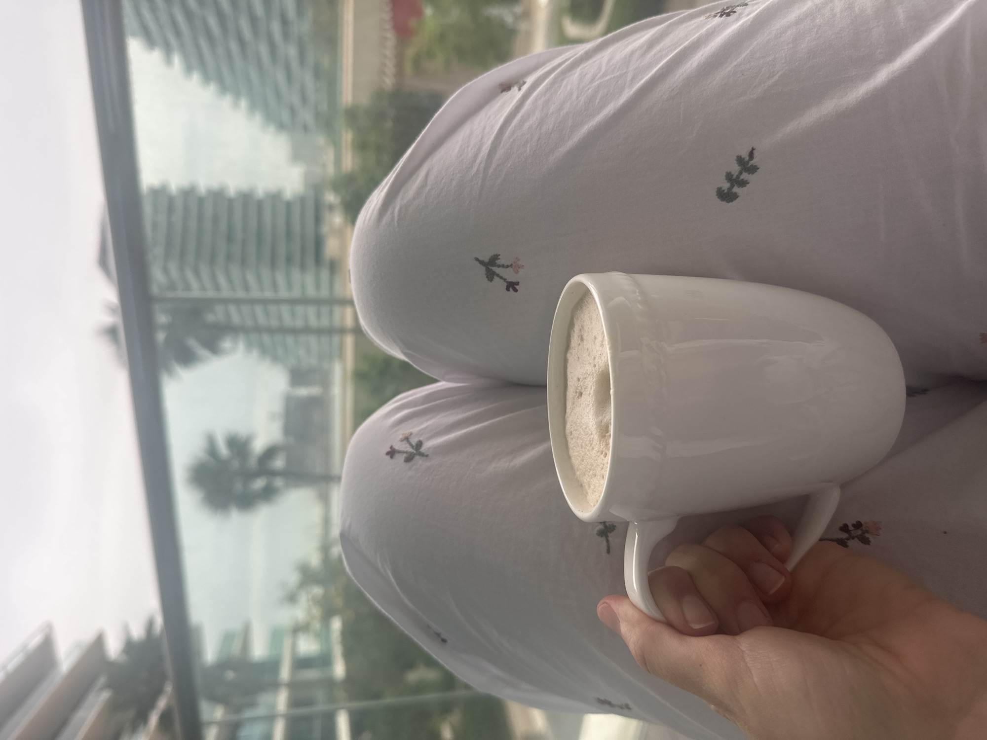A person sits with crossed legs, holding a white mug filled with a foamy beverage in their right hand. The person is wearing white pajama pants with small floral patterns. Outside a window, high-rise buildings and palm trees are visible, suggesting a city environment, likely Abu Dhabi, United Arab Emirates. The scene is taken indoors, possibly from a high vantage point, with the window offering a view of the city skyline.