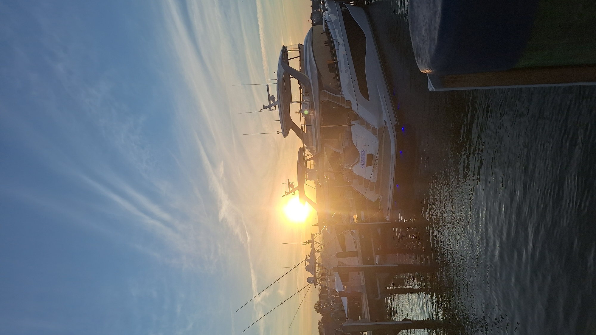 The image depicts a marina on Nantucket Island, Massachusetts, at sunset. The sun is low in the sky, casting a warm glow and creating lens flare. Several large yachts are docked, their sleek lines illuminated by the setting sun. One yacht in the foreground has the name "SALAMANDER" visible on its stern. Fishing rods are set up on smaller boats docked further back. The water is calm, reflecting the sky and the boats. The sky is a gradient of blue and orange, with wispy clouds streaking across it, characteristic of a sunset. The overall scene conveys a sense of peacefulness and luxury.