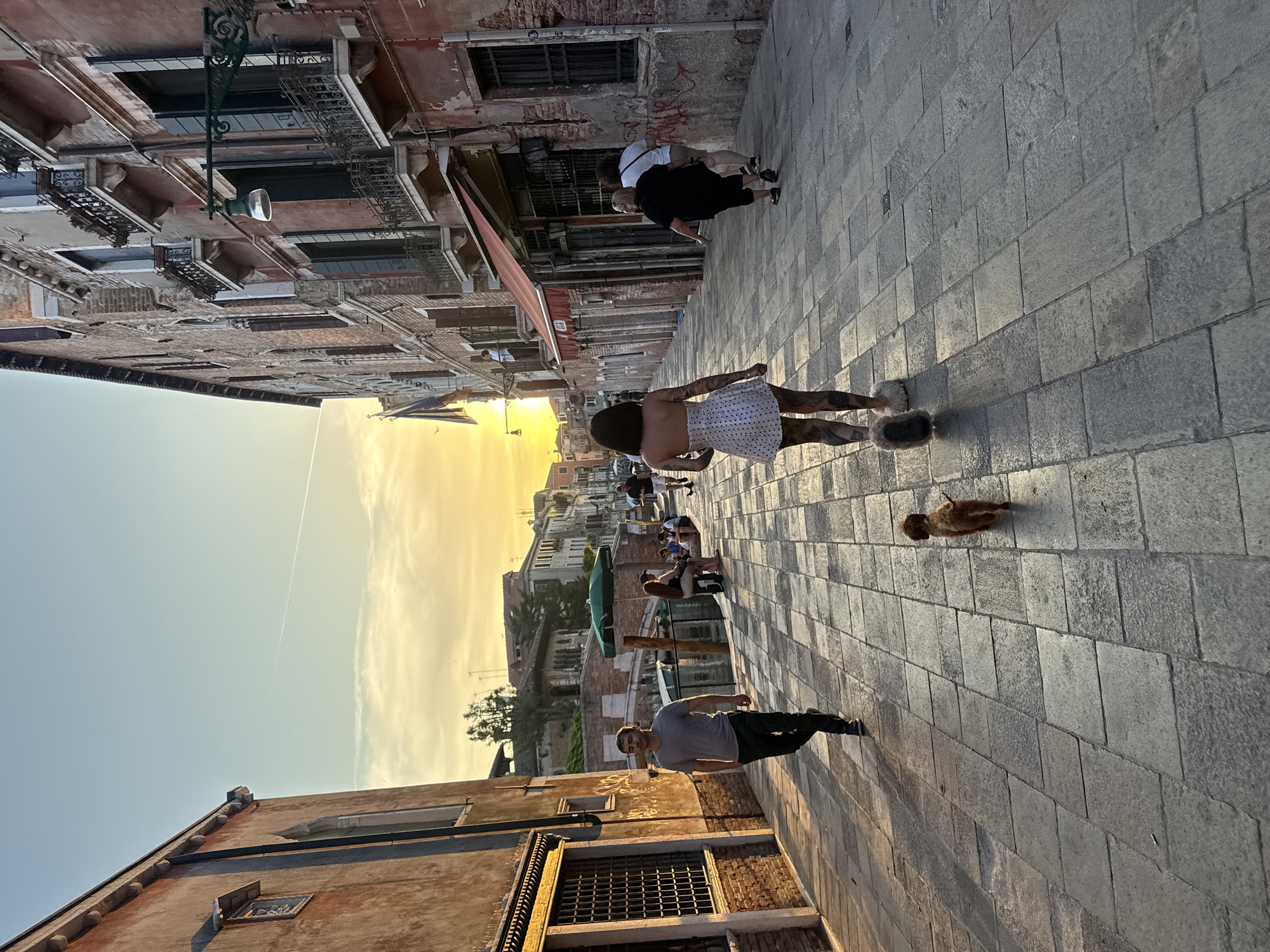 The image captures a vibrant street scene in Venice, Italy, during the golden hour of sunset. The perspective is from a slightly elevated vantage point, looking down a narrow calle (street) lined with classic Venetian buildings.
The sky in the distance glows with warm orange and yellow hues where the sun is setting, transitioning to a softer blue overhead. A faint contrail streaks across the clear sky. The buildings on both sides of the street are old, with weathered brick and stucco facades in earthy red, brown, and grey tones. Many windows feature dark frames and ornate iron railings. A prominent lamppost with a modern cylindrical light fixture extends from one building on the left side.
The street itself is paved with large, rectangular grey stone blocks, showing signs of age and wear. Several people are walking along the street. In the foreground, a man in a grey t-shirt and dark pants walks towards the viewer. Behind him, a woman with dark hair, wearing a white polka-dot dress and distinct furry slippers, walks away, revealing tattooed legs. Further back, two other figures, one in a white top and another in a black top, also walk away. In the distance, near what appears to be a canal, more people are visible, some seated near a green outdoor umbrella.
On the paved ground, a small, brown duck-like bird can be seen. The low sun casts long, distinct shadows of the people and buildings across the street, highlighting the texture of the paving stones. The overall atmosphere is tranquil and leisurely, typical of an evening stroll in a historic city. No legible text or signs are visible.