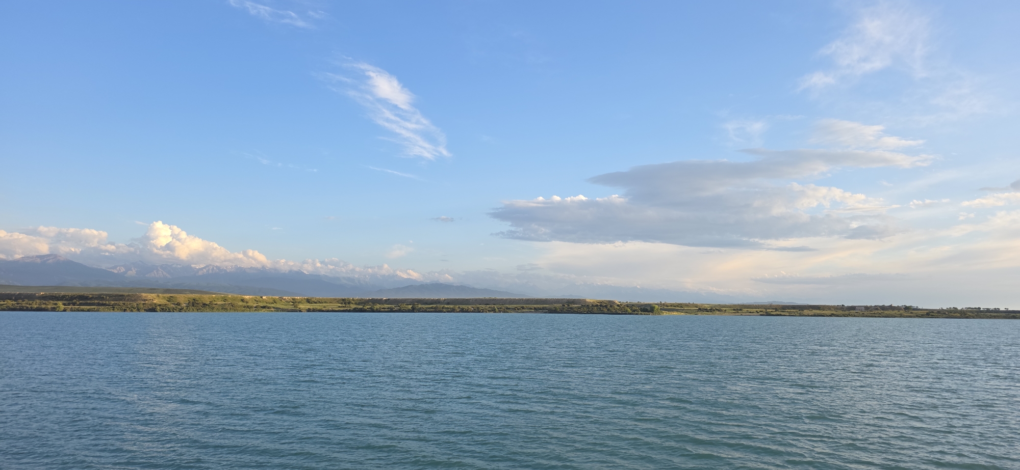 This is a scenic, wide-angle, landscape photograph taken from the shore of a large body of water, likely Issyk-Kul Lake, near Pristan'-Przheval'sk, Kyrgyzstan. The foreground is dominated by the rippling, turquoise surface of the lake, reflecting the bright blue sky. The horizon line divides the image, with the distant shore featuring a strip of green land dotted with sparse vegetation and some scattered buildings. Beyond the land, a majestic mountain range stretches across the background, its peaks capped with snow and illuminated by the soft light of the setting sun. Wispy white clouds drift across the expansive sky, adding depth and texture to the scene. The overall impression is one of tranquility and natural beauty. There are no people or animals visible in the image.
