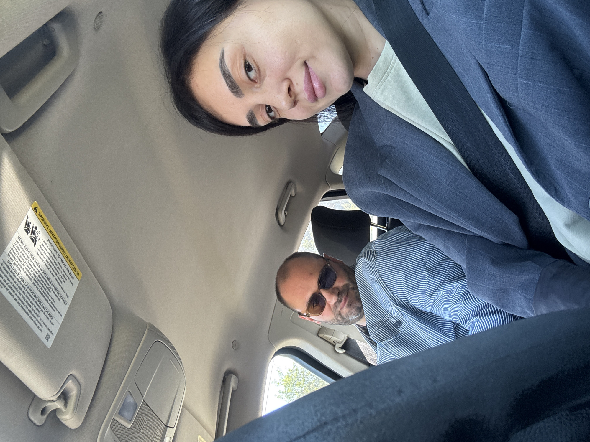 The image shows two people inside a car. A woman is facing the camera from the front passenger side, wearing a grey blazer and a seatbelt. A man with a shaved head, sunglasses, and a striped shirt is in the back. The interior of the car is visible, including the dashboard, sun visor, and headliner. The car is likely in motion. The location is not discernible from the image.