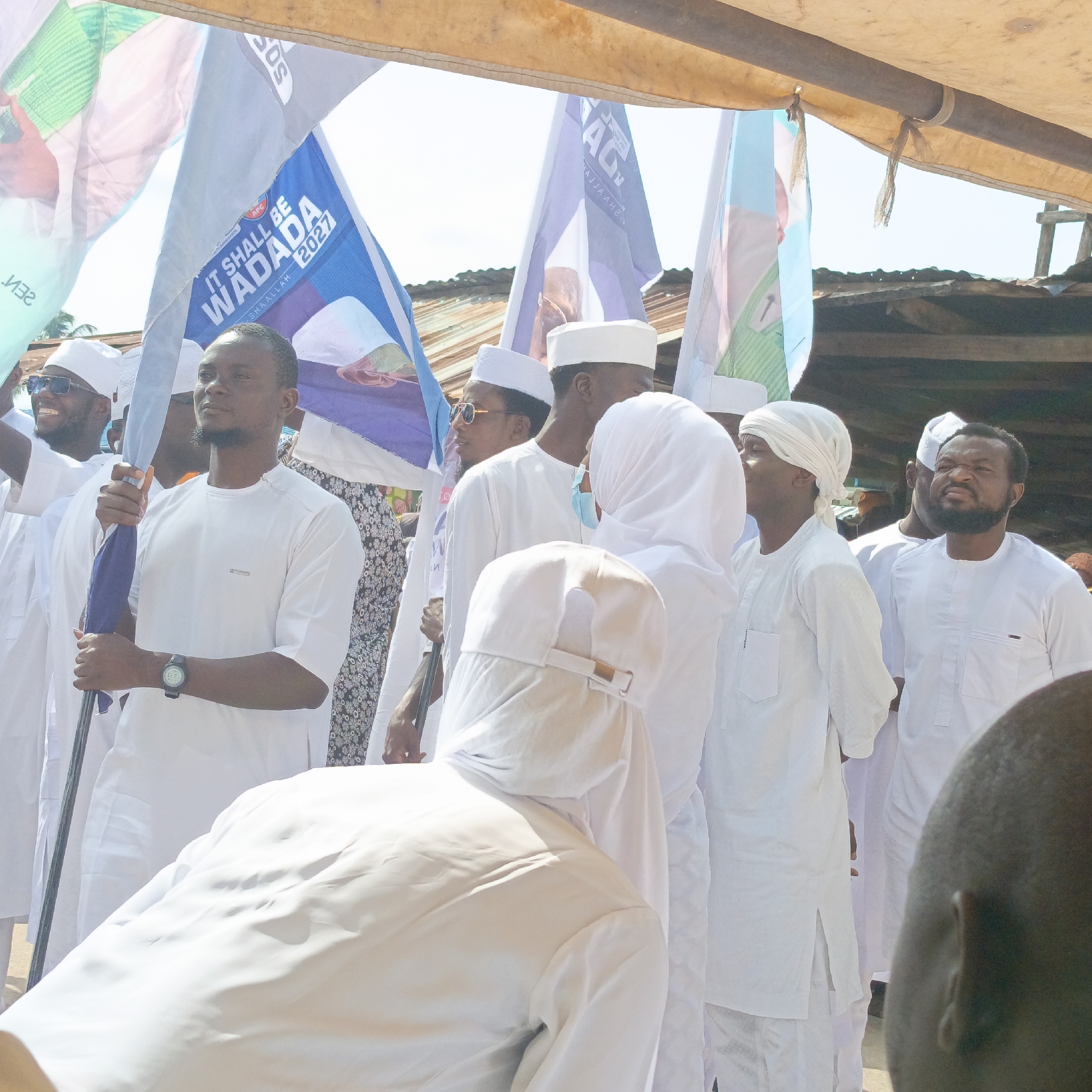The image captures a procession in Akwanga, Nigeria, where a group of men are walking together. Most are dressed in white traditional outfits, including white caps. One man is holding a flag with the text "IT SHALL BE WADADA 2027." Other flags are visible as well. The men appear to be involved in a political or celebratory event. The background includes a metal roof of a building and a makeshift awning providing shade.